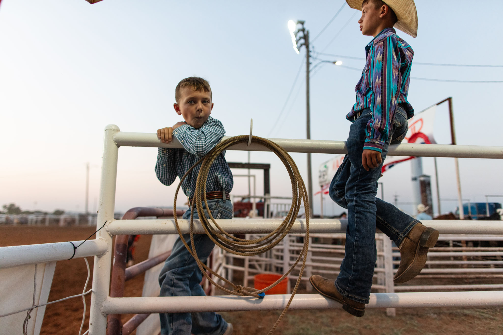 Two boys sitting on the arena fence, one holding a rope