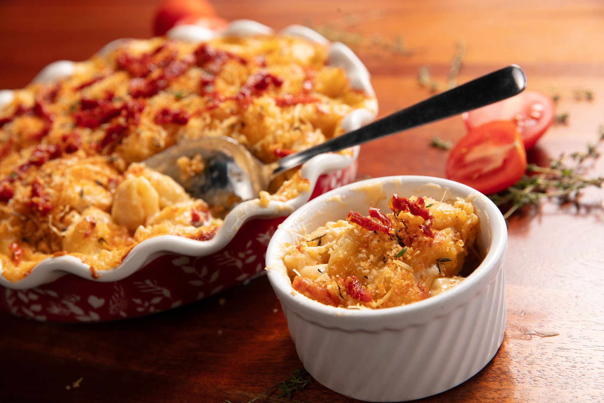 Baked macaroni and cheese with a golden breadcrumb crust in a ruffled serving dish and a white ramekin, with a spoon, thyme and tomato halves