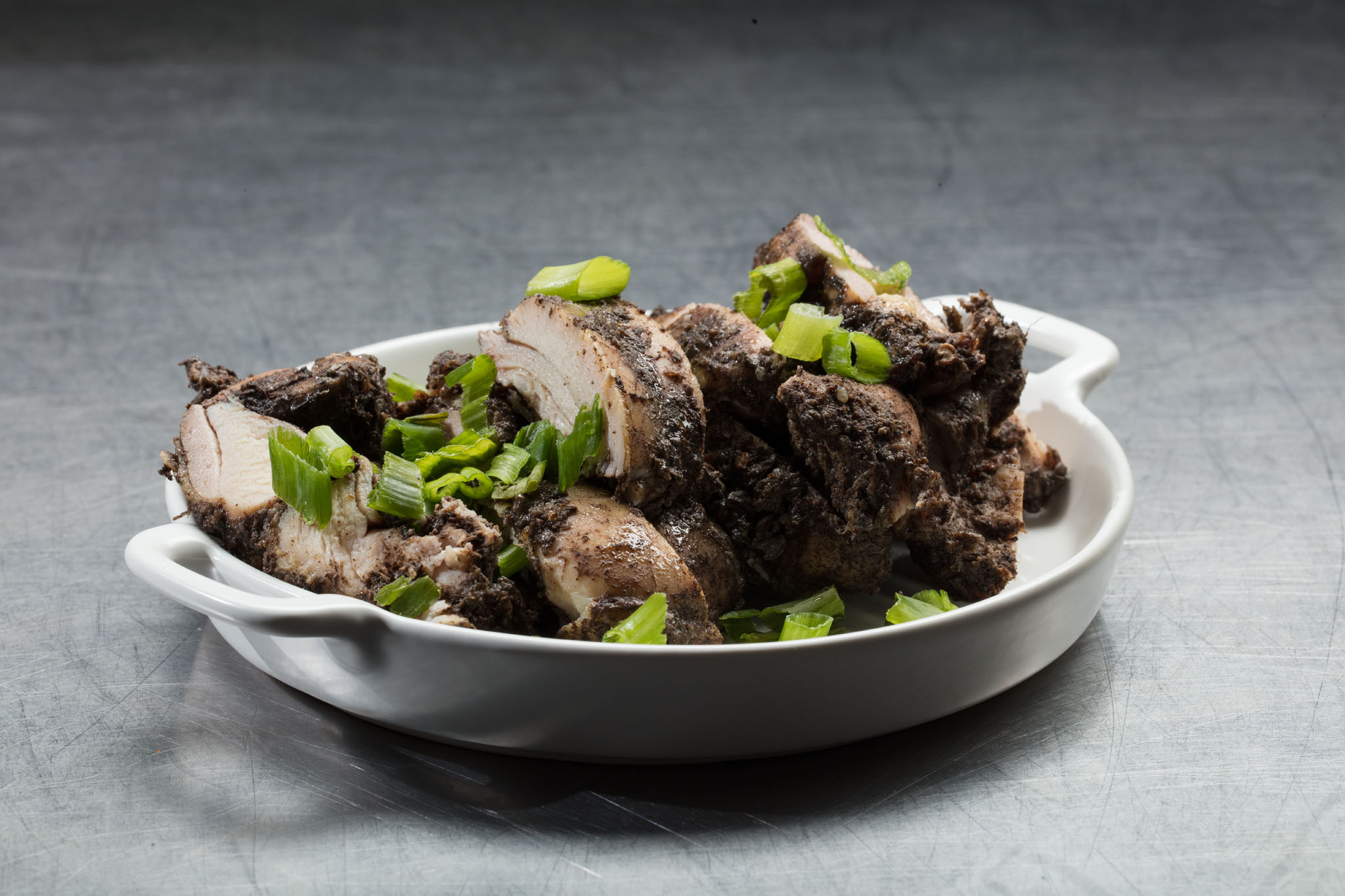 Sliced chicken coated in a dark seasoning and garnished with green onion in a white oval dish on a gray textured surface