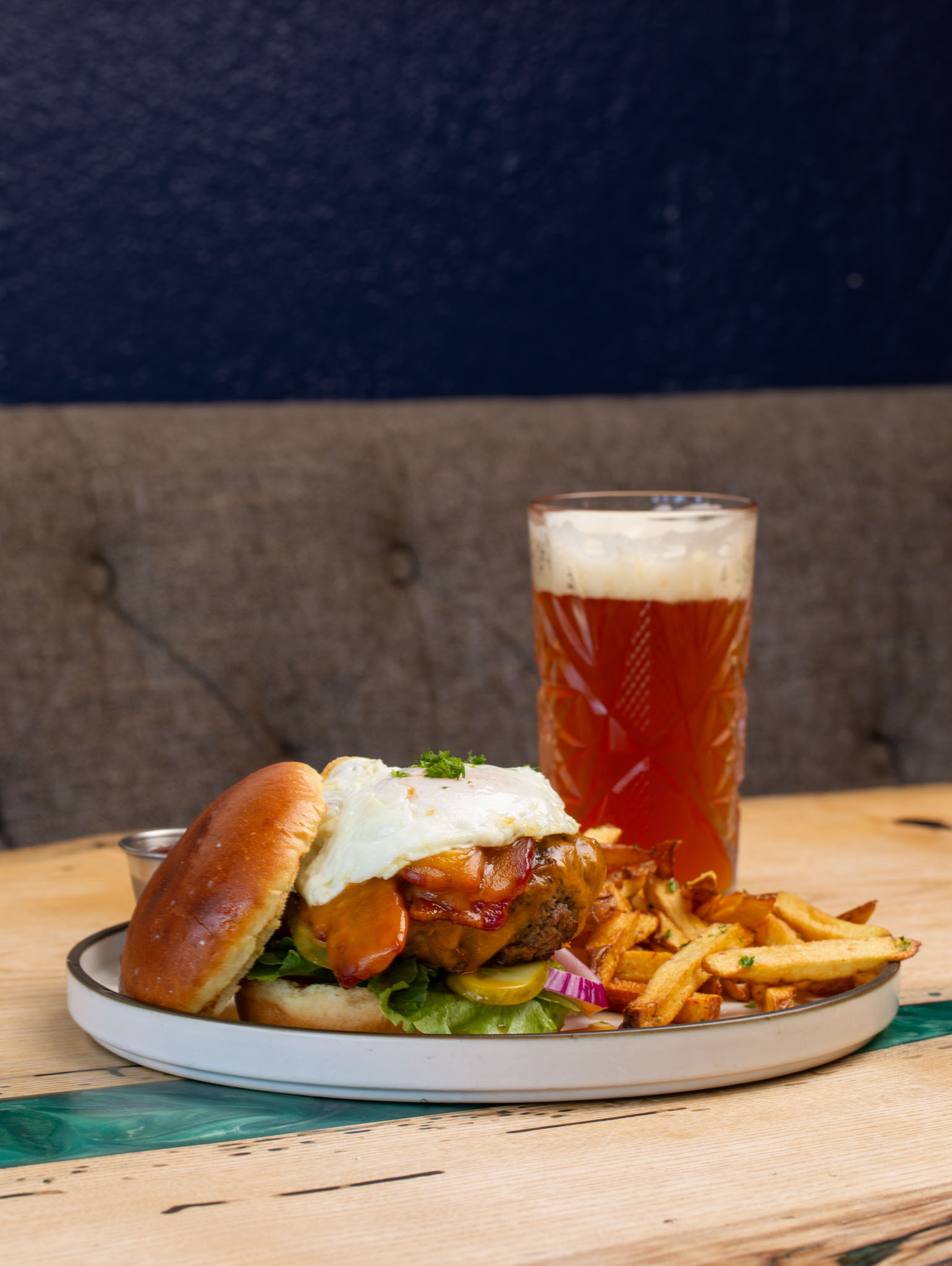 Burger topped with a fried egg and served with fries on a plate beside a tall glass of amber beer, against a tufted booth