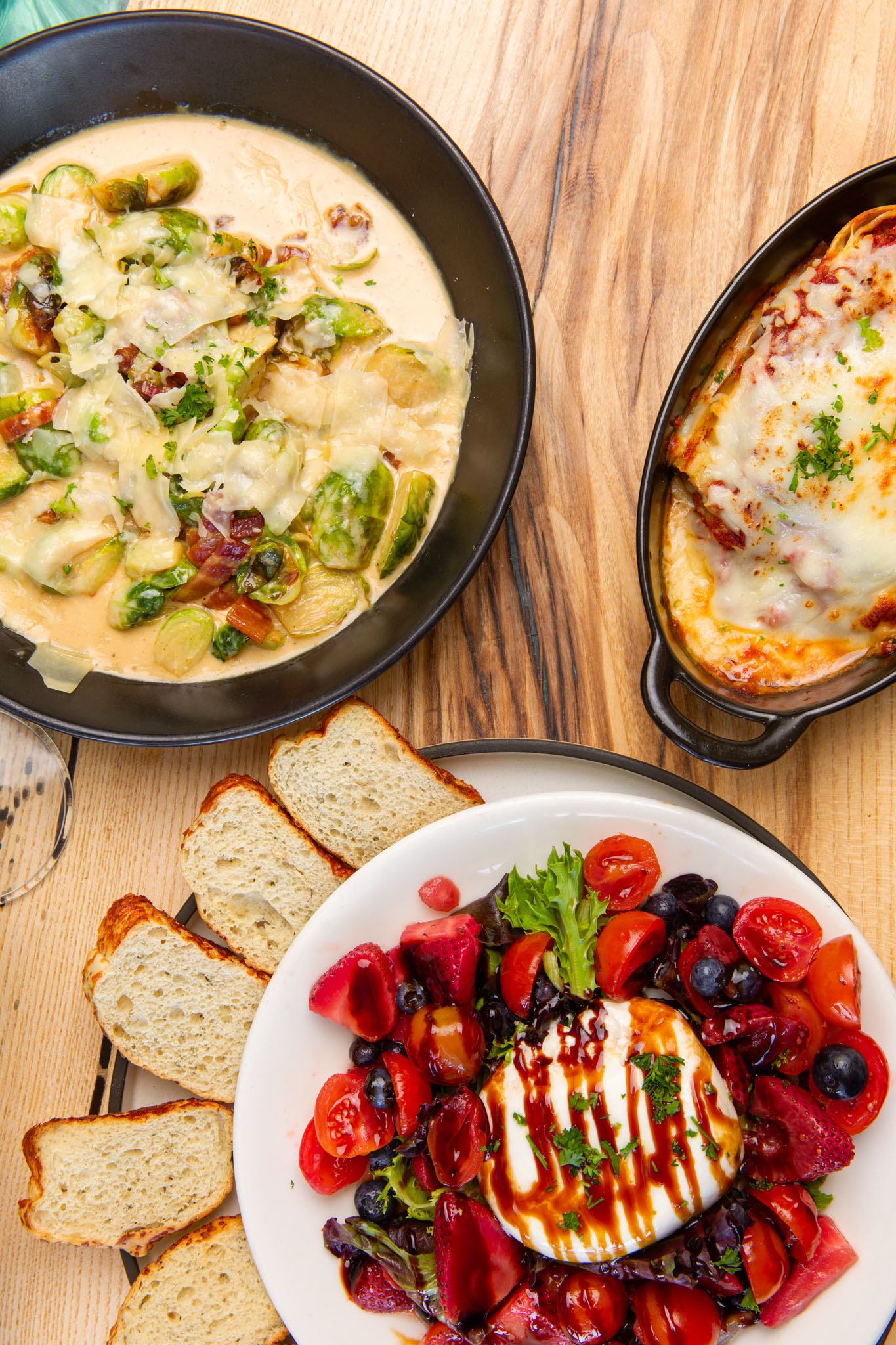Overhead view of a table spread with creamy Brussels sprouts in a skillet, a cheese-topped baked dish, sliced bread and a burrata salad with tomatoes and berries