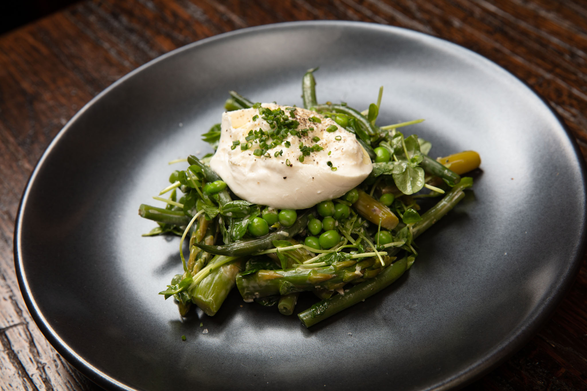 Spring vegetable salad of green beans, asparagus, peas and pea shoots topped with burrata and chives on a dark plate