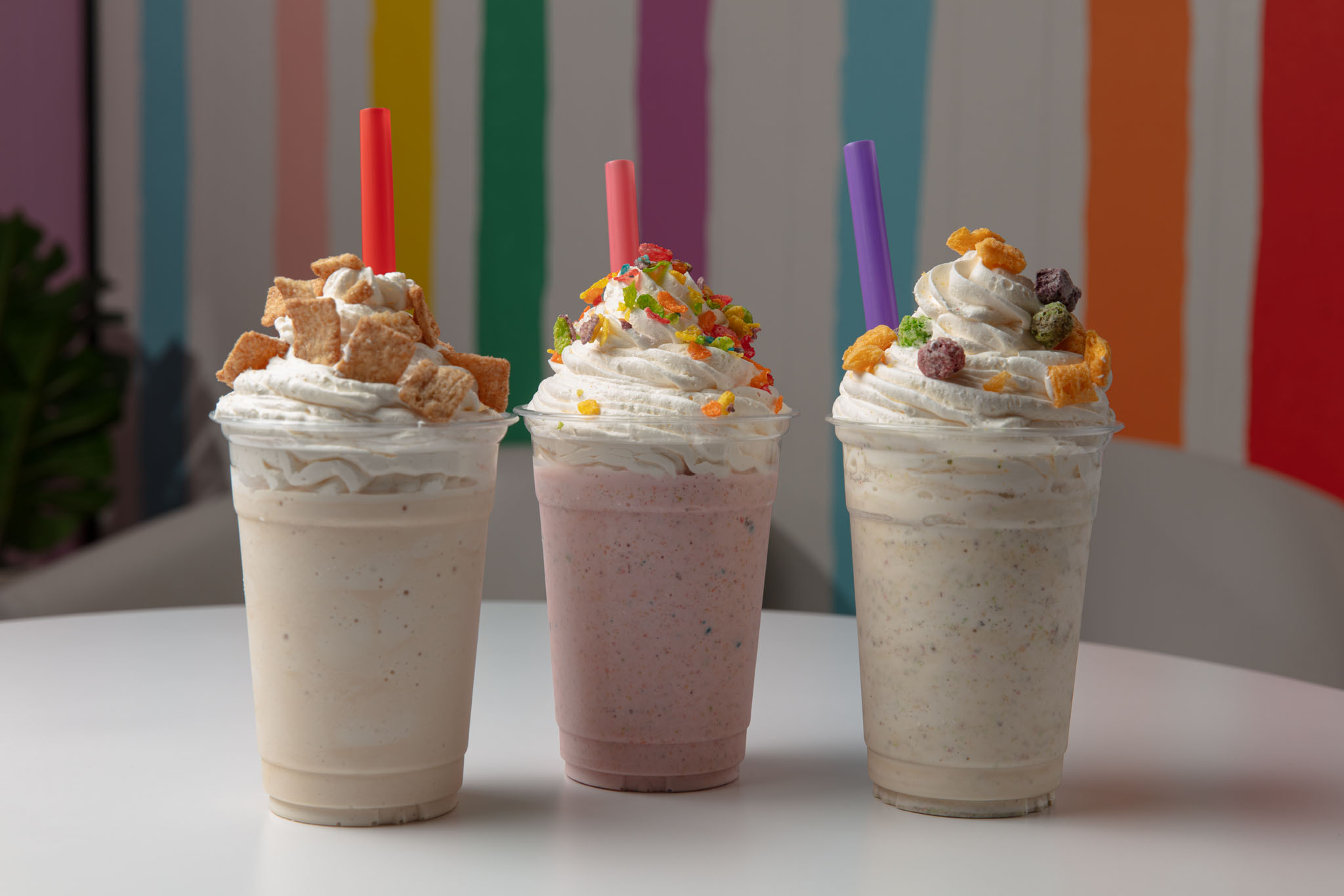Three milkshakes topped with whipped cream and cereal with colorful straws, in front of a wall with painted rainbow stripes