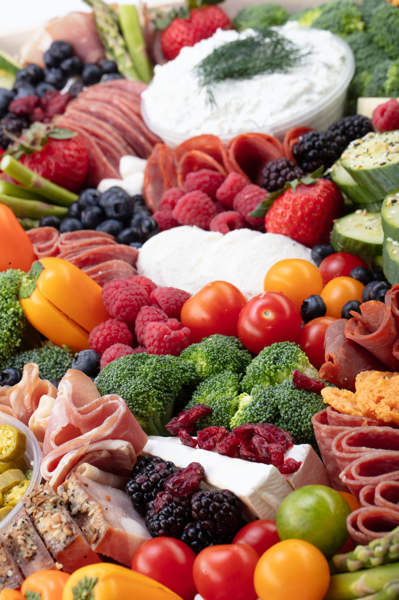 Overflowing grazing board with fresh berries, tomatoes, broccoli, cucumber, cured meats, cheeses and a bowl of dip garnished with dill