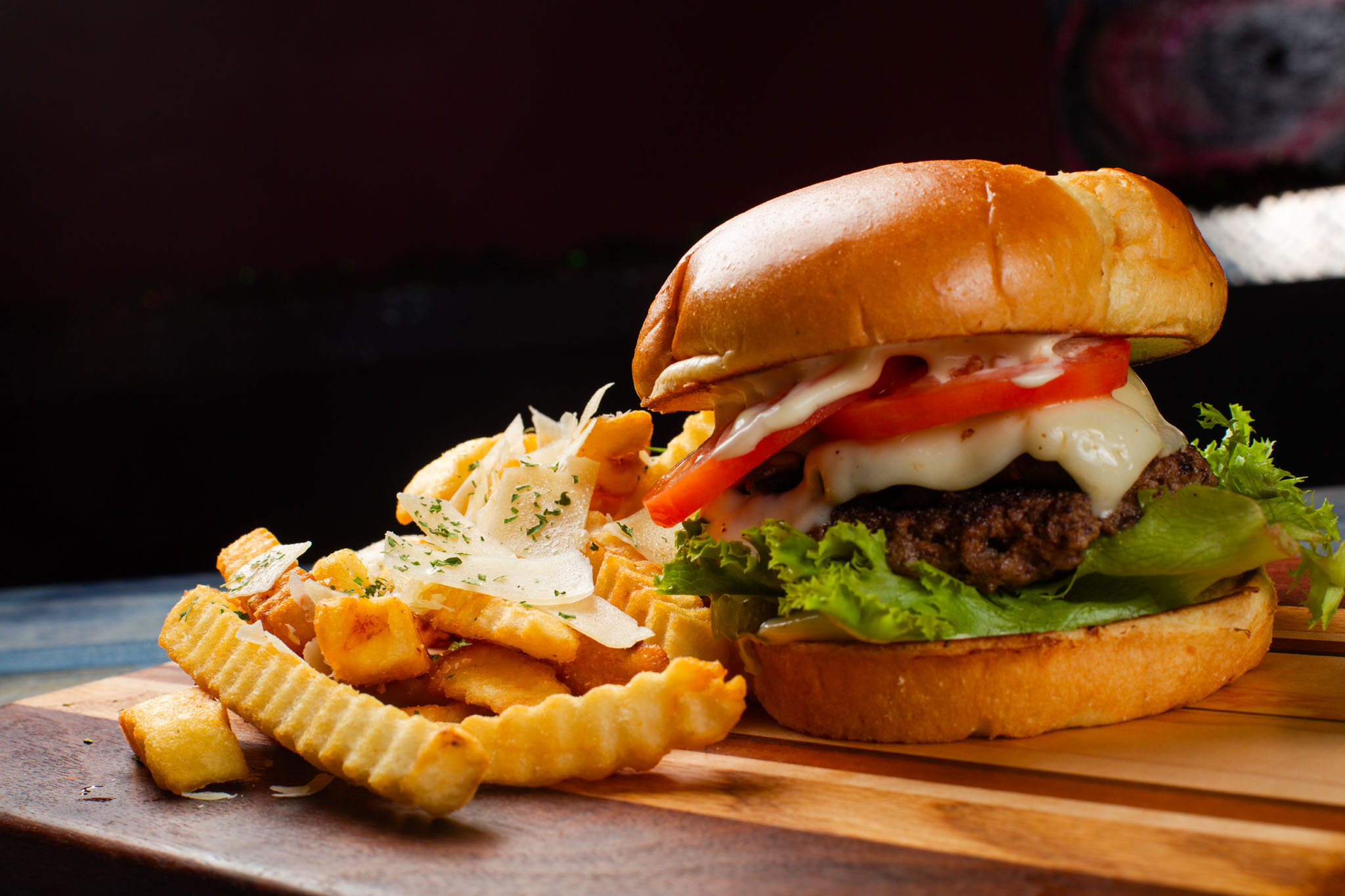 Cheeseburger with lettuce and tomato on a toasted bun beside crinkle-cut fries topped with shaved cheese and herbs, on a wooden board