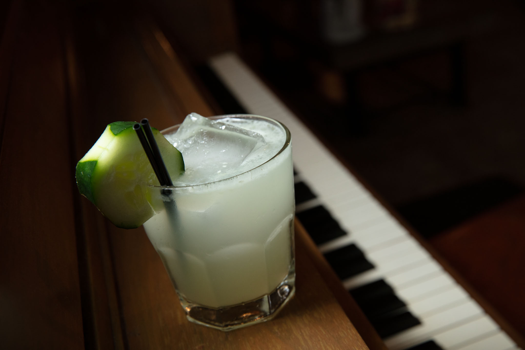 The Cucumber cocktail at Saints, a pale green drink over ice with a cucumber garnish and black straw, set on a piano