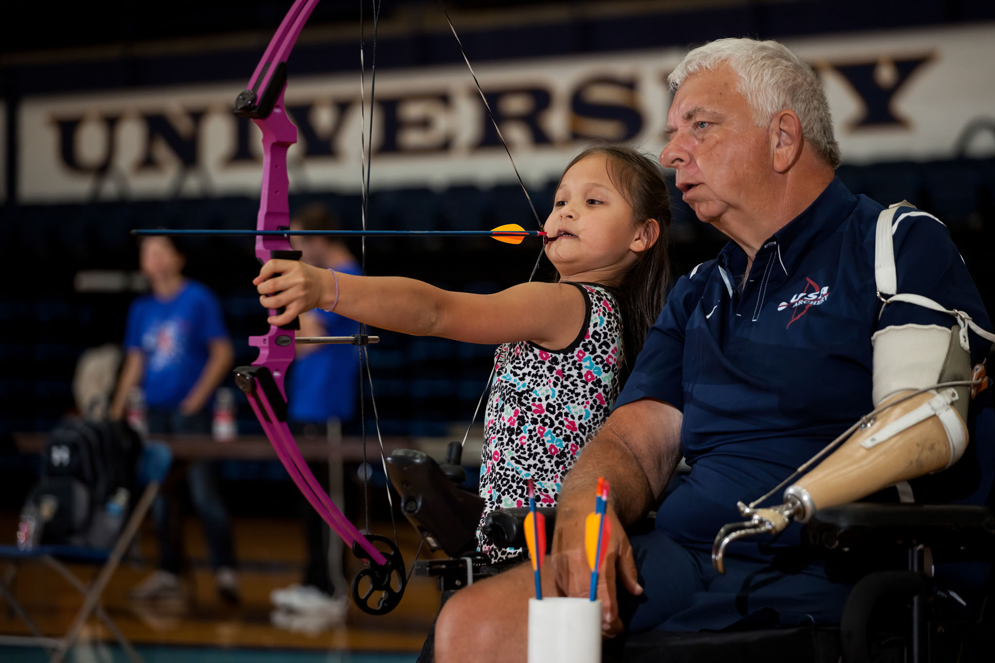 Young girl drawing a pink bow beside a seated coach at an archery range