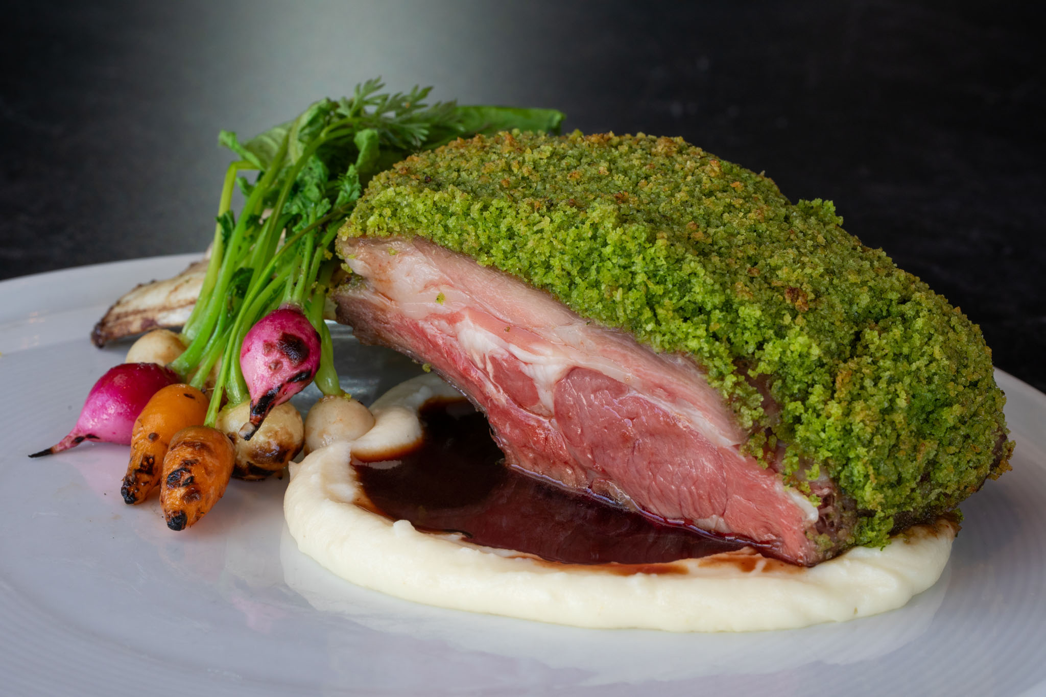 Herb-crusted roast with a bright green crust served over puree and jus with charred baby carrots and radishes