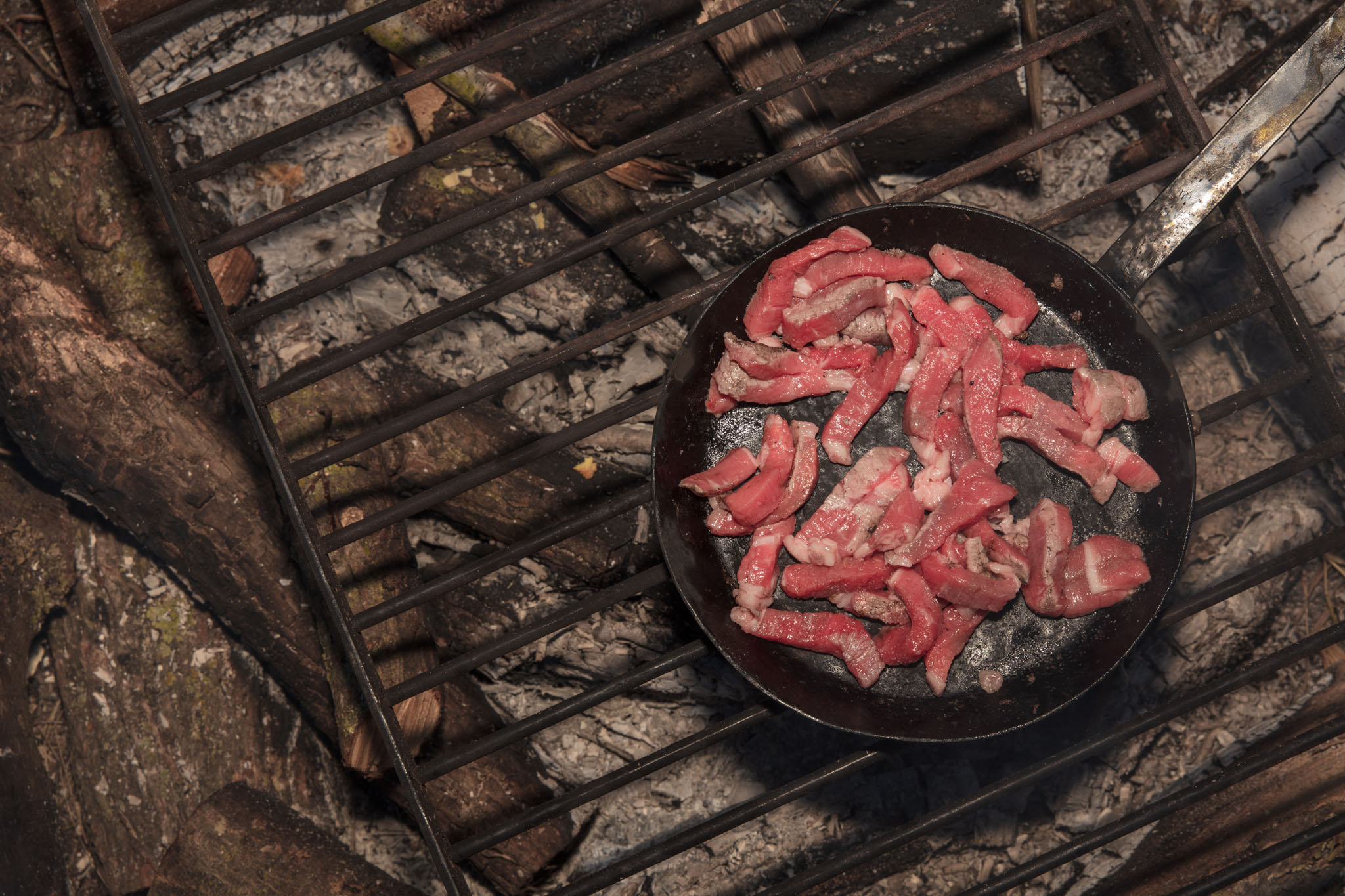 Bacon frying in a cast-iron skillet over a campfire grate