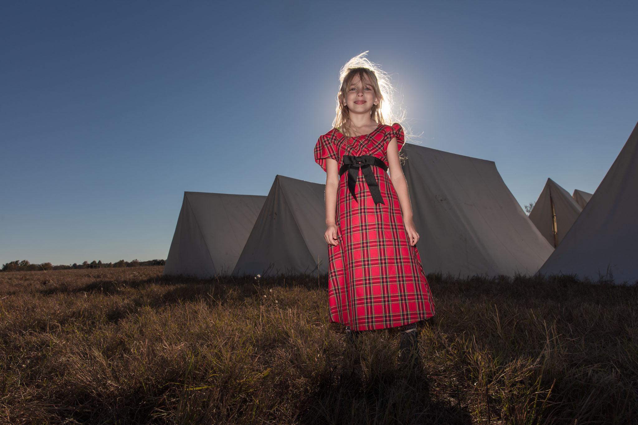 Girl in a red plaid dress standing before the tents with the sun behind her