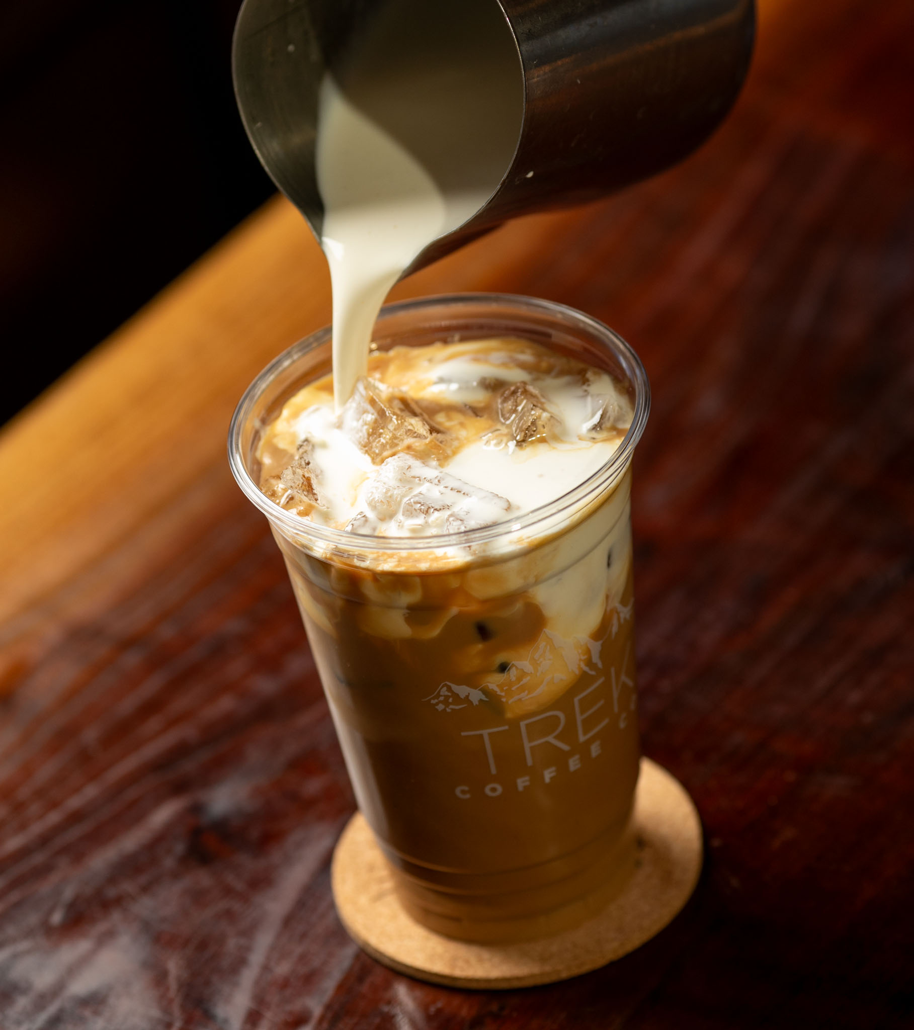Milk being poured into an iced coffee in a Trek Coffee cup on a dark wood table
