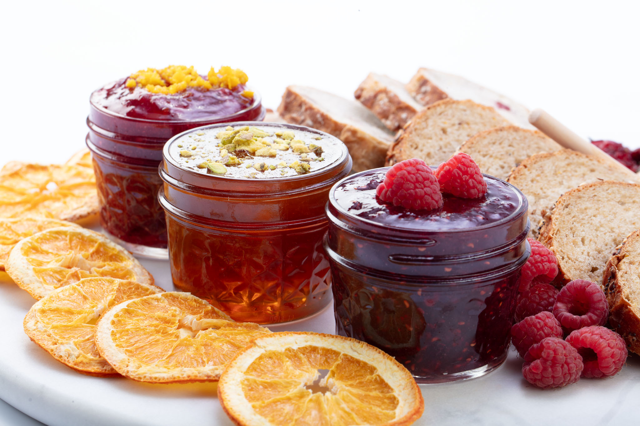Three jars of fruit preserves with fresh raspberries, dried orange slices and sliced bread on a white board