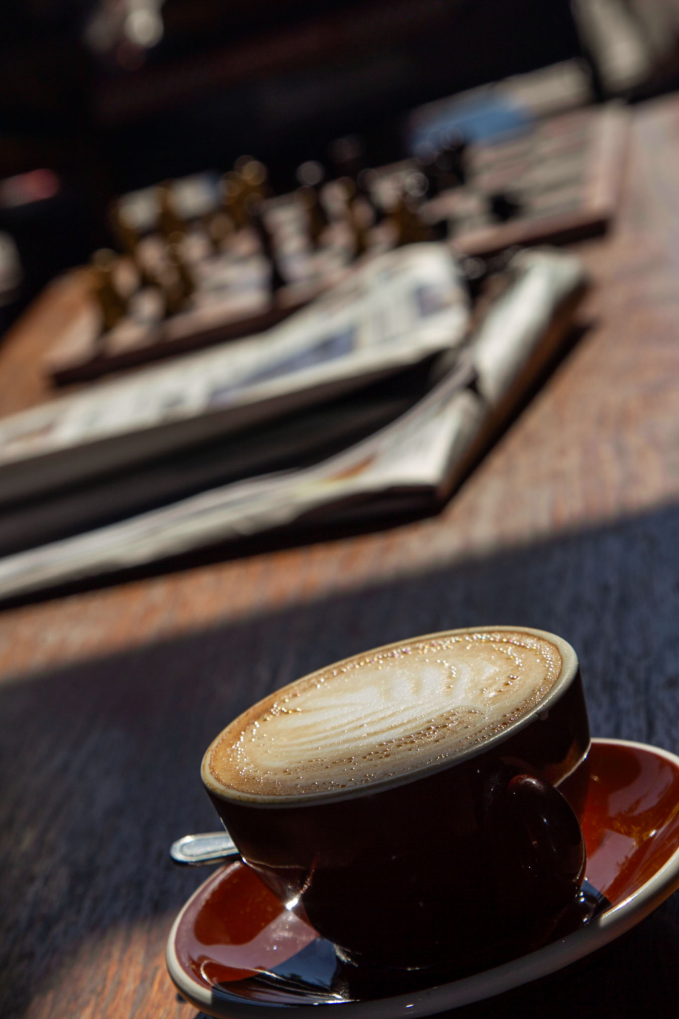 Cappuccino with latte art in a dark cup on a red saucer on a wooden table in window light, with folded newspapers blurred behind