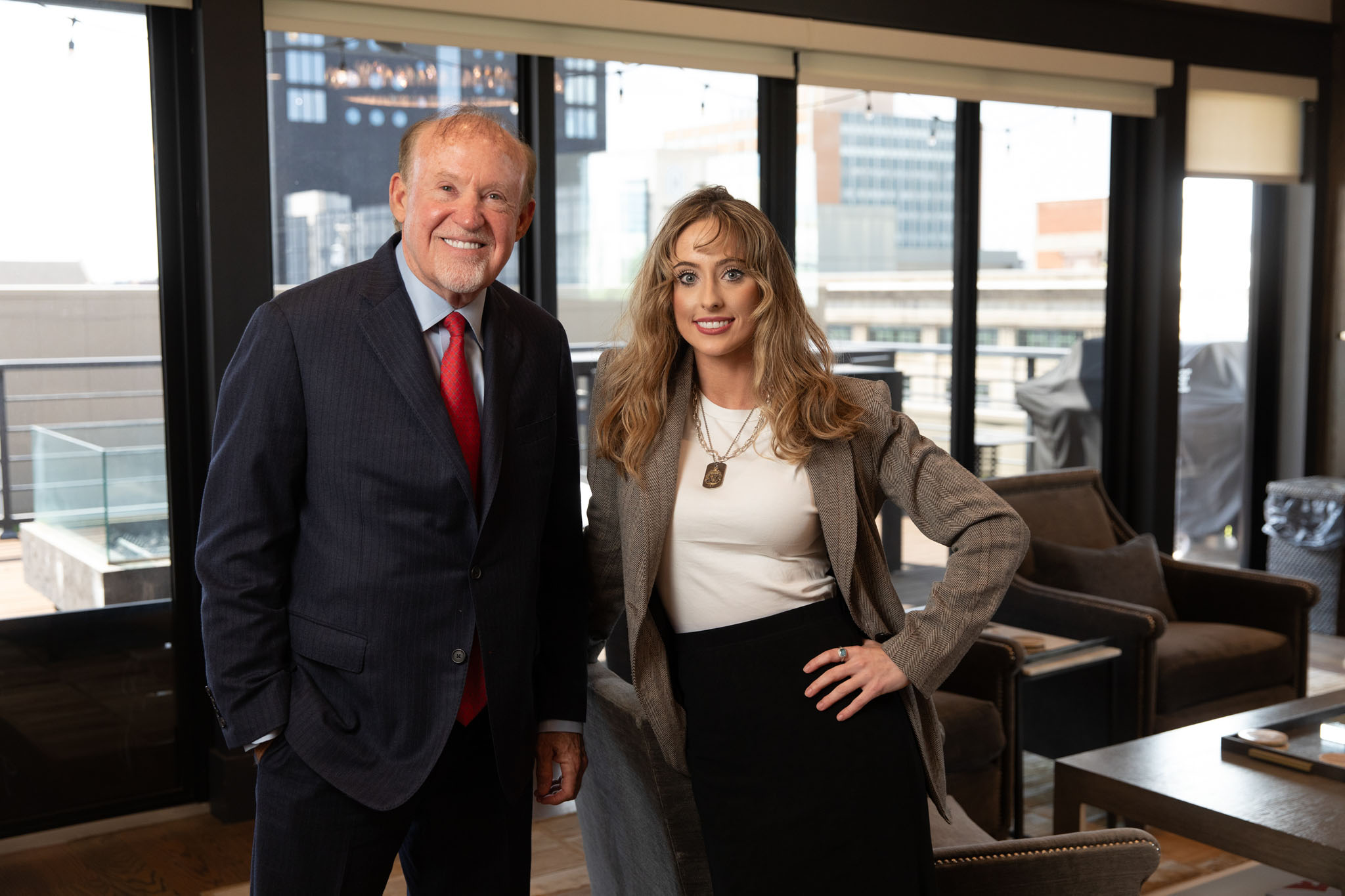 Man in a navy suit and red tie standing with a woman in a tweed blazer in an office with city windows