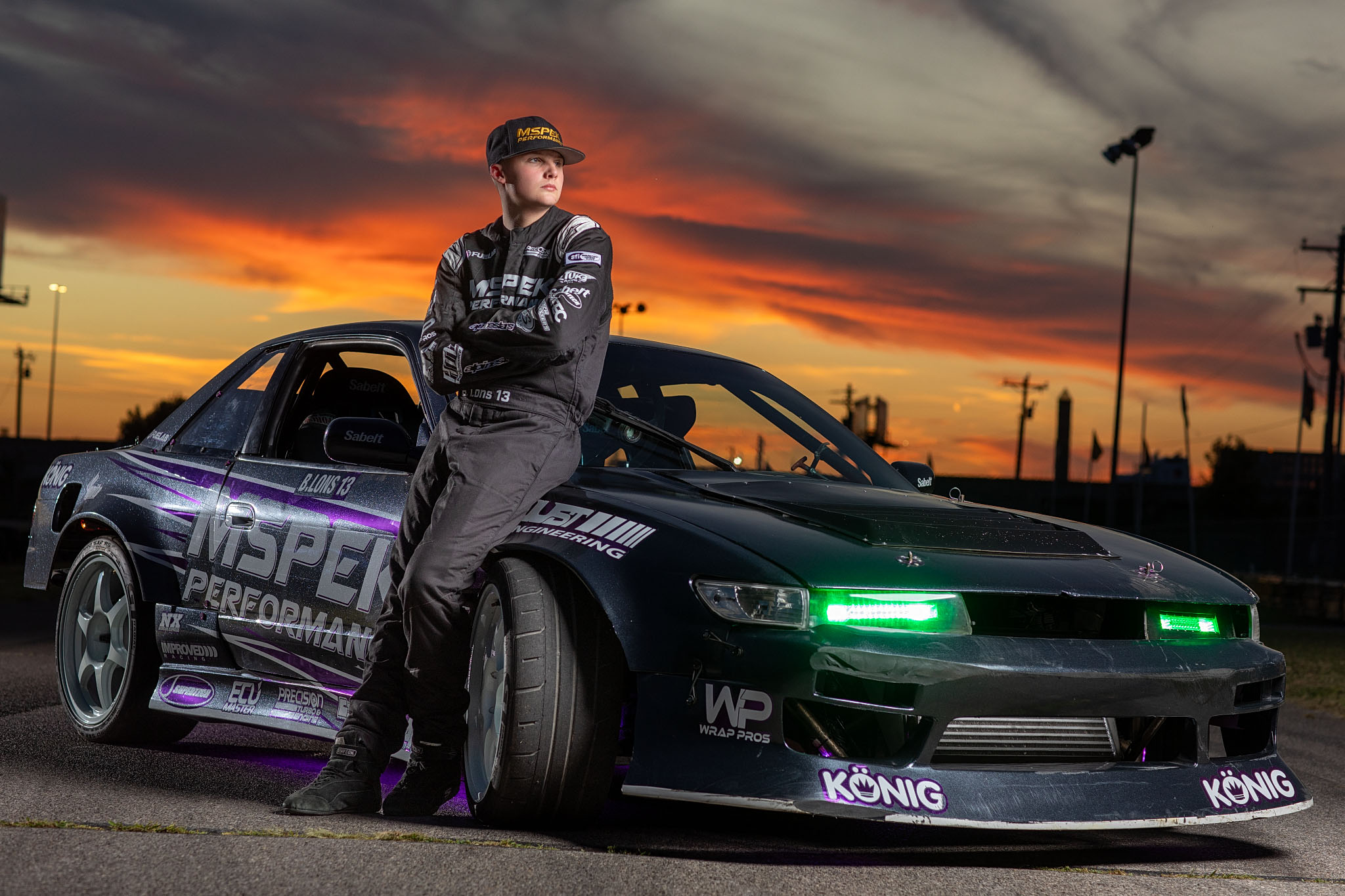 Race car driver in a fire suit leaning on a drift car at sunset