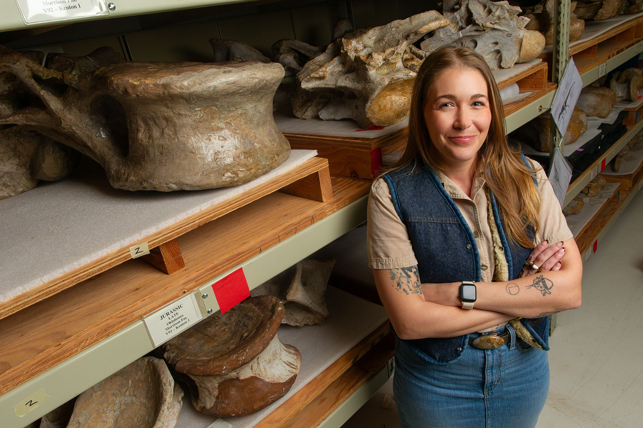Woman in a denim vest standing with arms crossed among museum storage shelves of artifacts