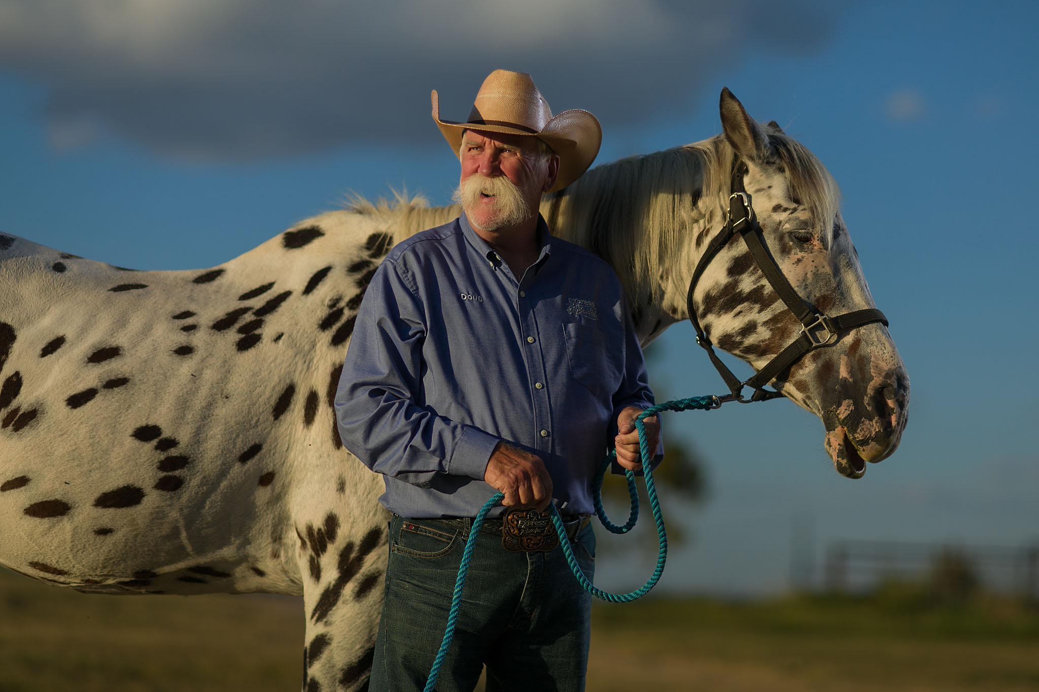 Man in a cowboy hat standing with a spotted Appaloosa horse in a pasture