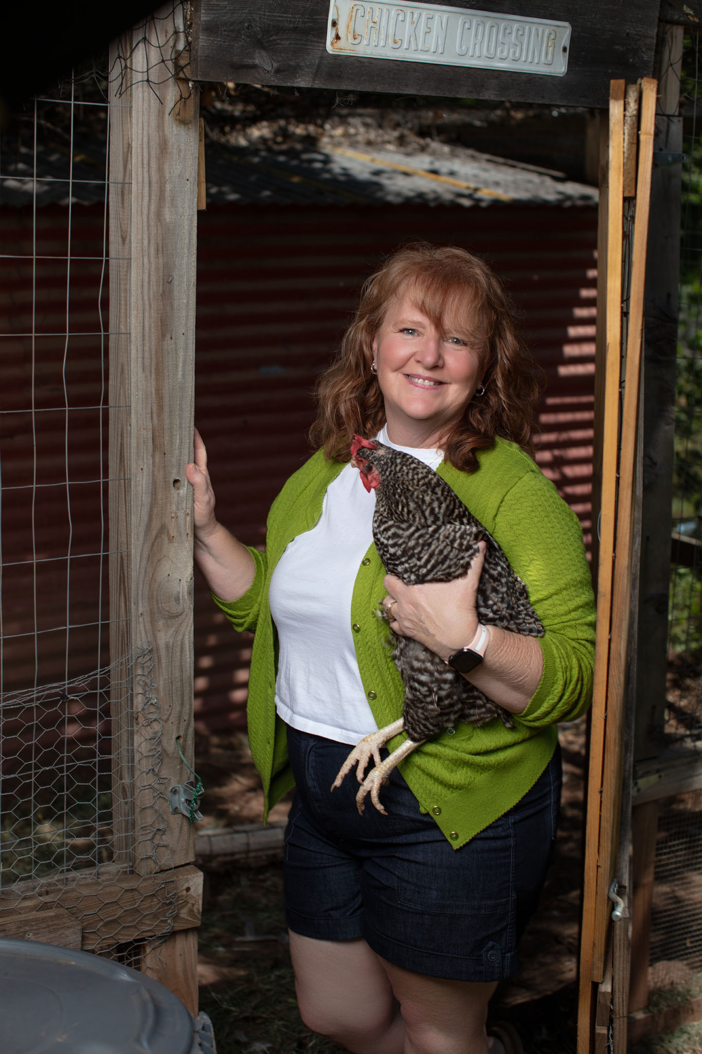 Smiling woman in a green cardigan holding a chicken in the doorway of a wooden chicken coop