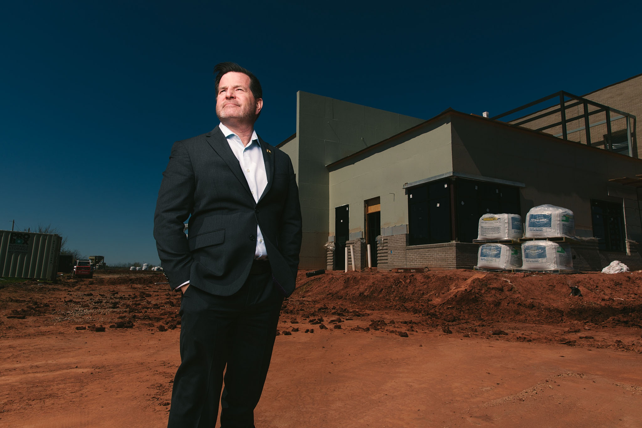 Man in a dark suit standing on red dirt in front of a new industrial building at dusk