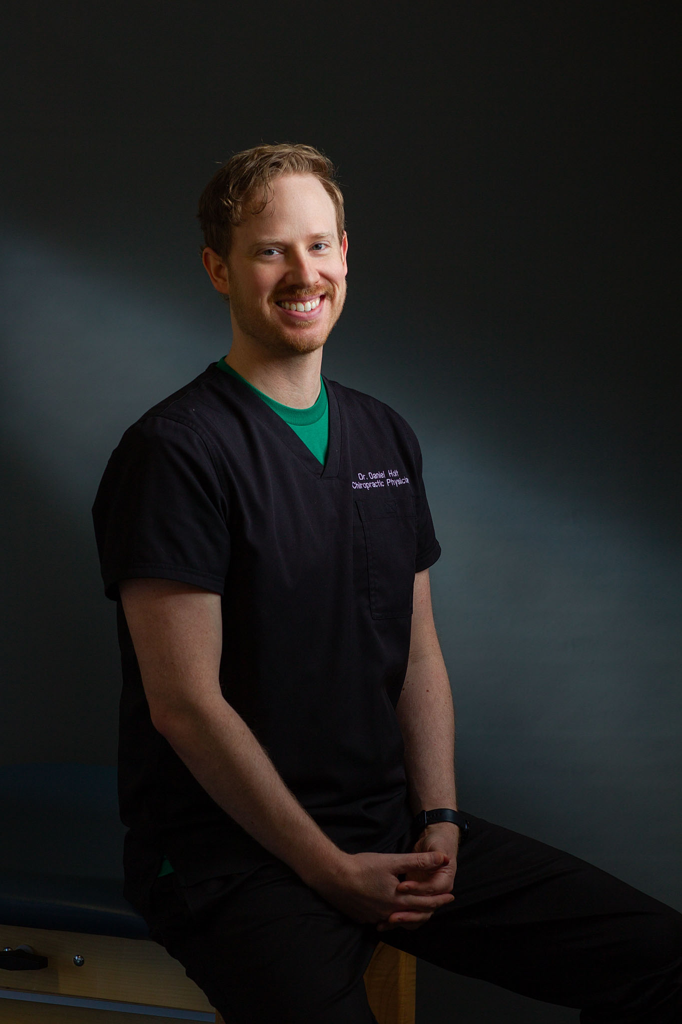 Smiling man in navy scrubs seated against a dark gray backdrop