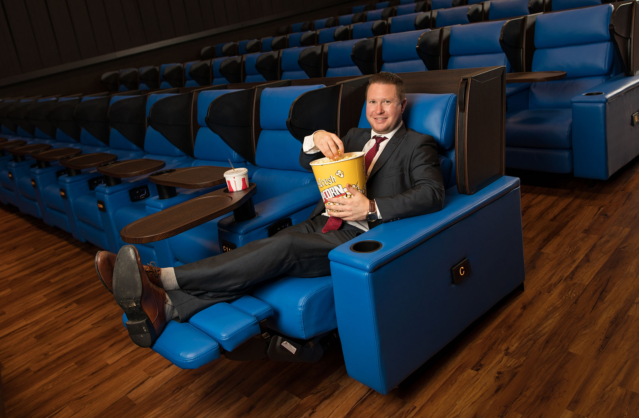 Man in a suit reclining with a bucket of popcorn in a blue leather theater recliner