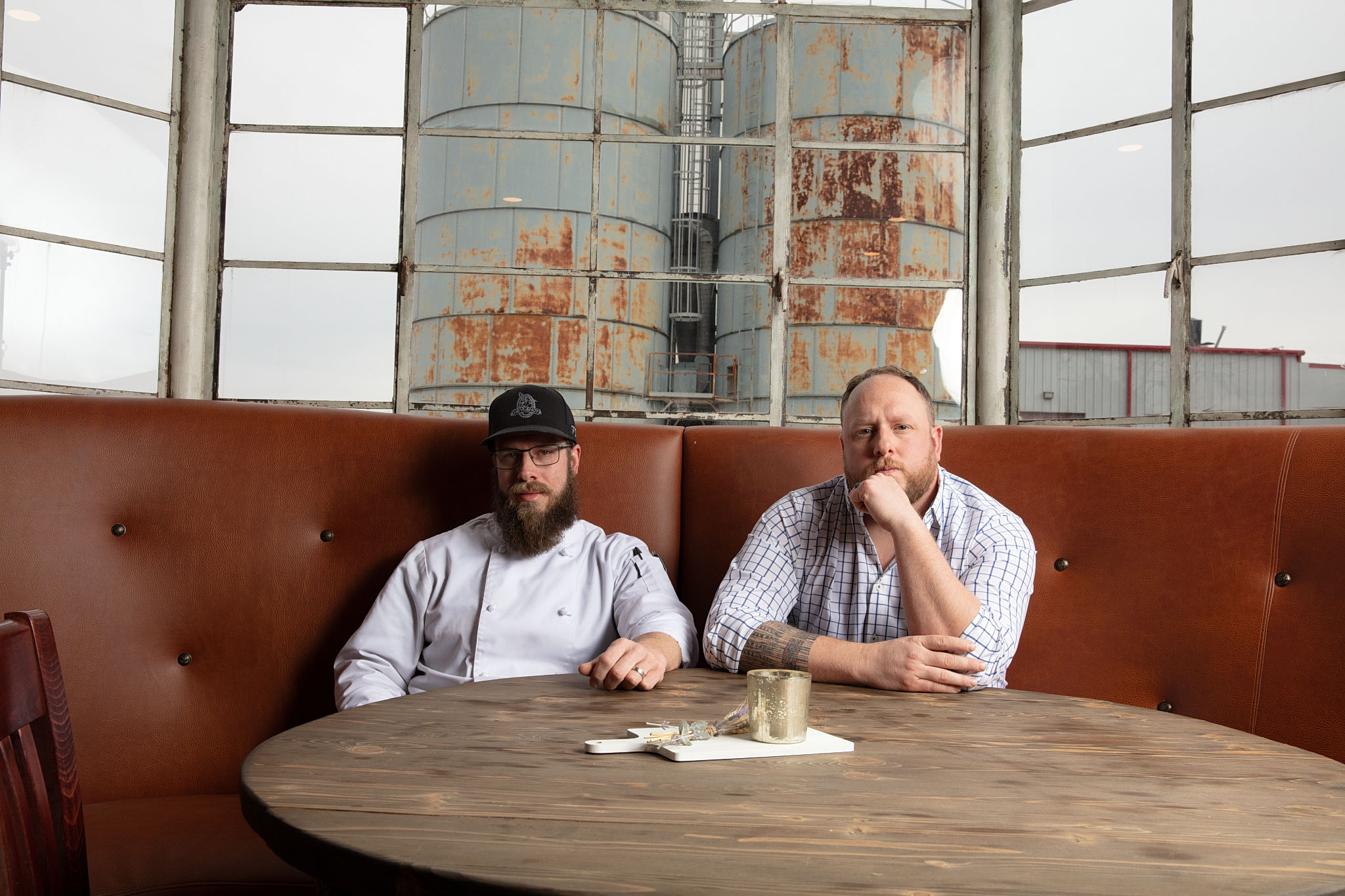 Two men seated in a leather booth at a round table in a restaurant with tall industrial windows