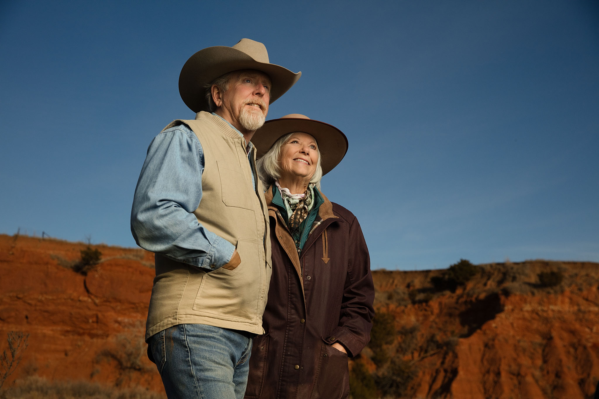 Older ranch couple in cowboy hats standing above red canyon land