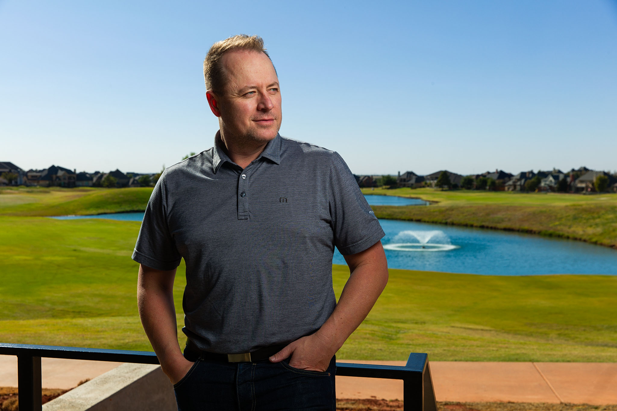 Man in a gray polo standing on a patio overlooking a golf course pond