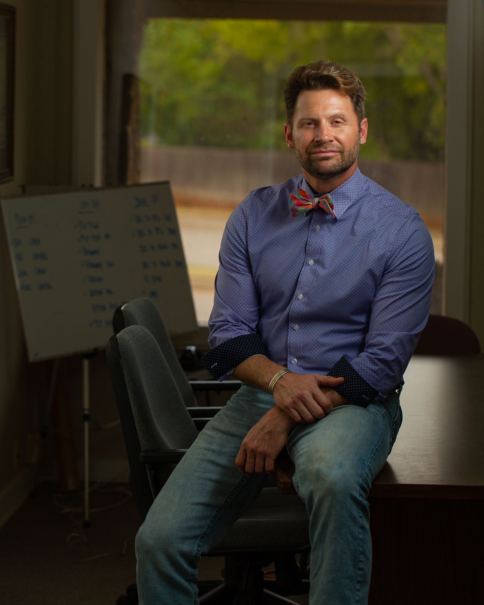 Man in a bow tie seated in front of a whiteboard in an office