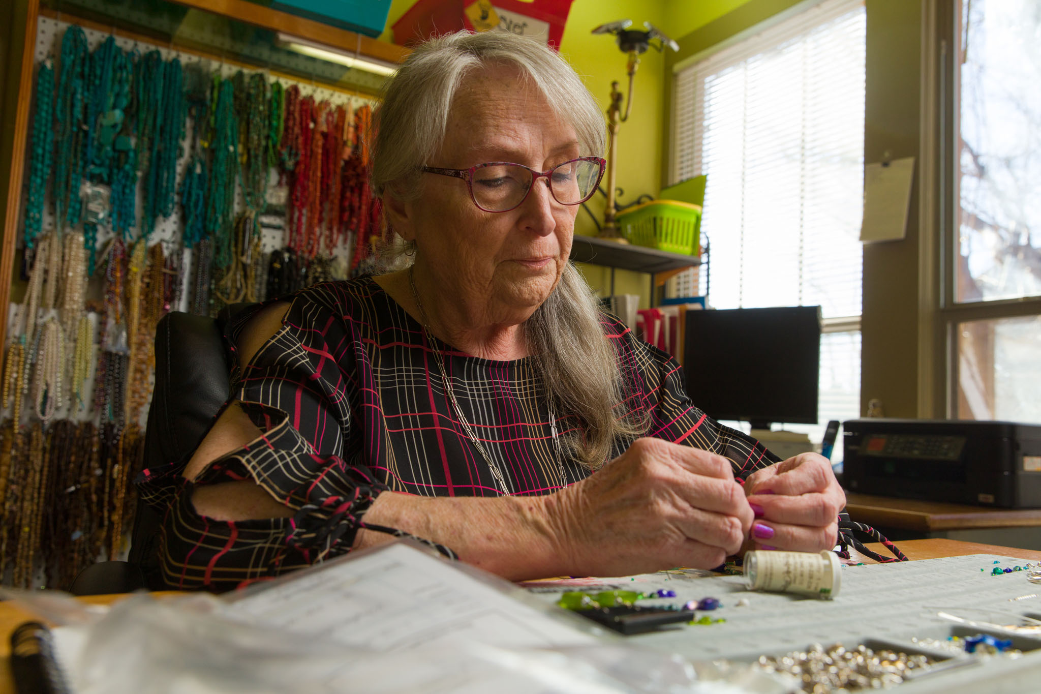 Woman with silver hair making beaded jewelry at a worktable surrounded by strands of beads