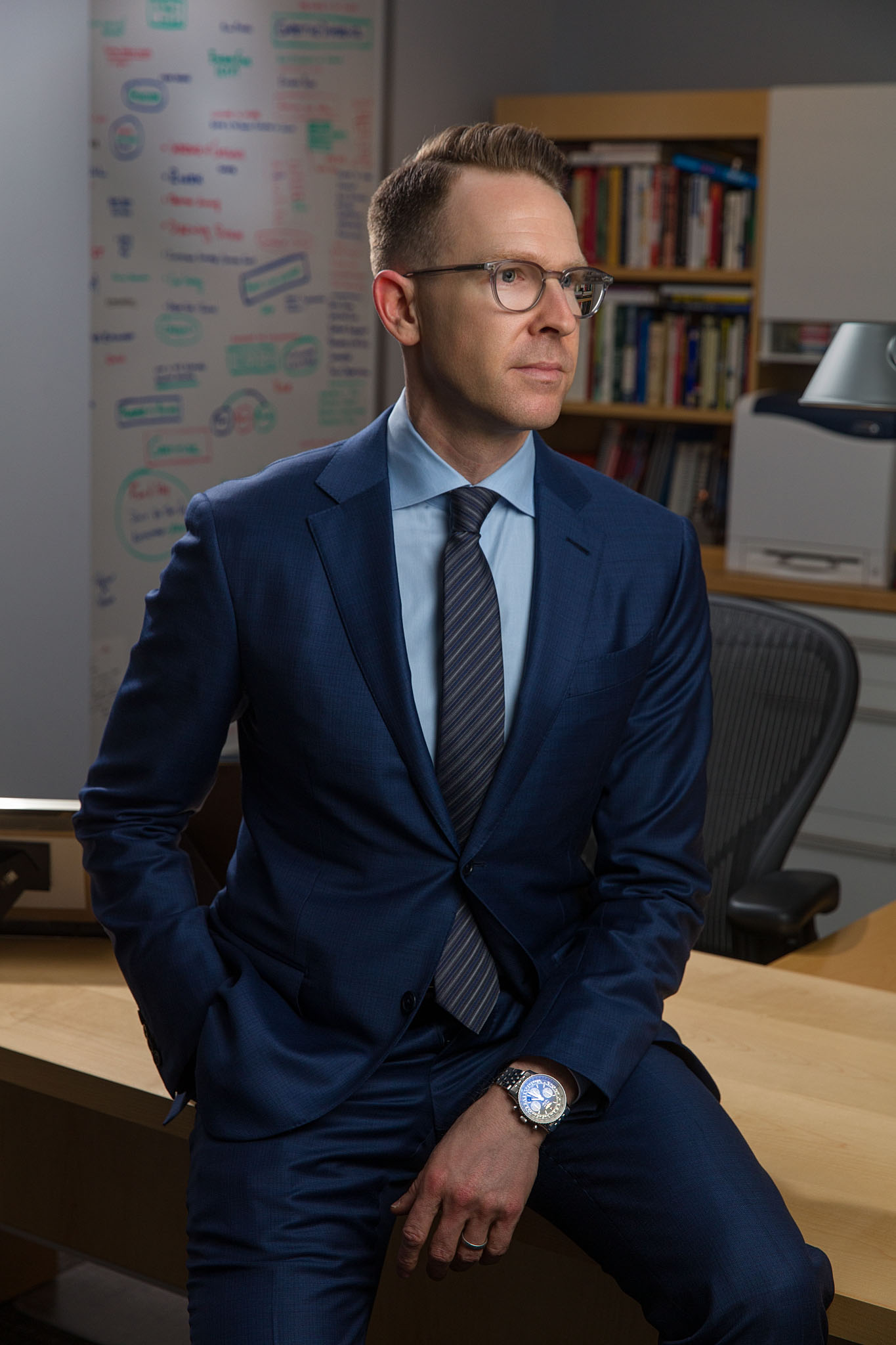 Man in glasses and a navy suit seated at a desk in an office with a whiteboard