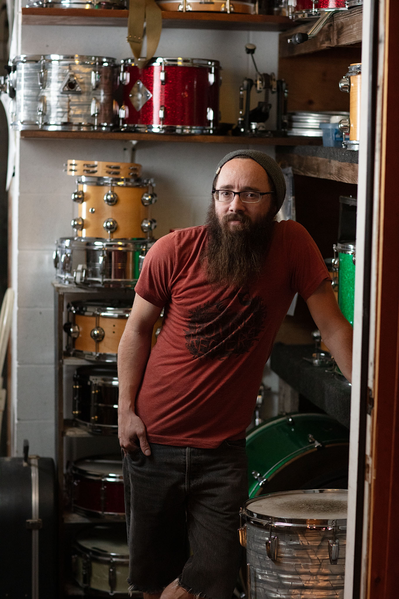 Bearded drummer in a red T-shirt standing among stacked drum kits