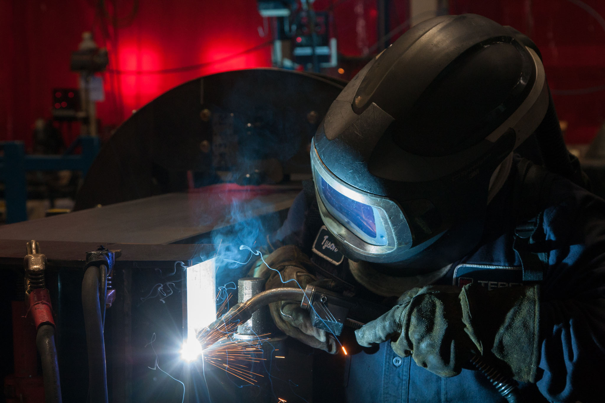 Welder in a helmet lit by a blue arc working under red light in a dark shop