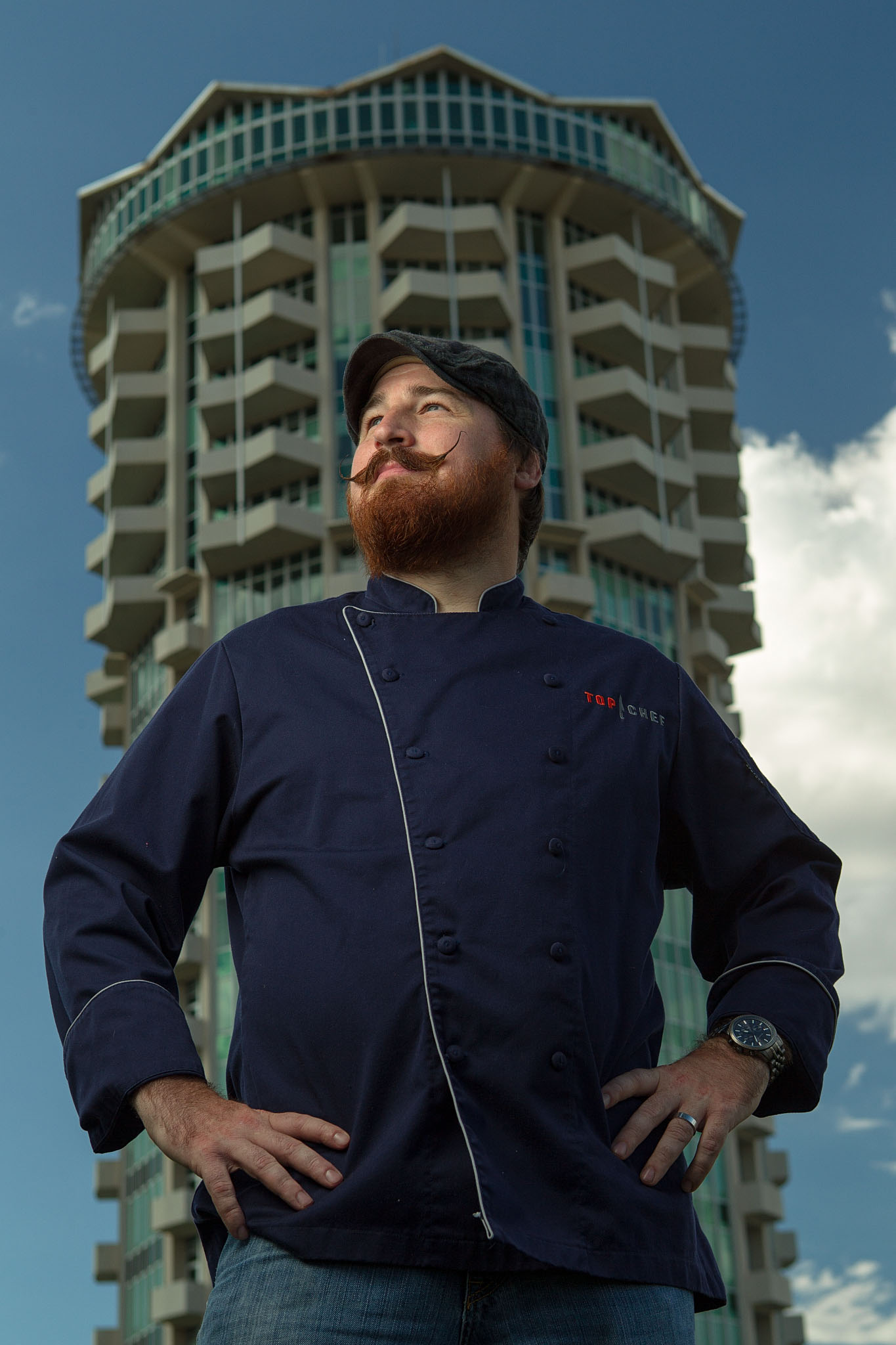Chef in a navy chef coat standing with hands on hips in front of a round tower building