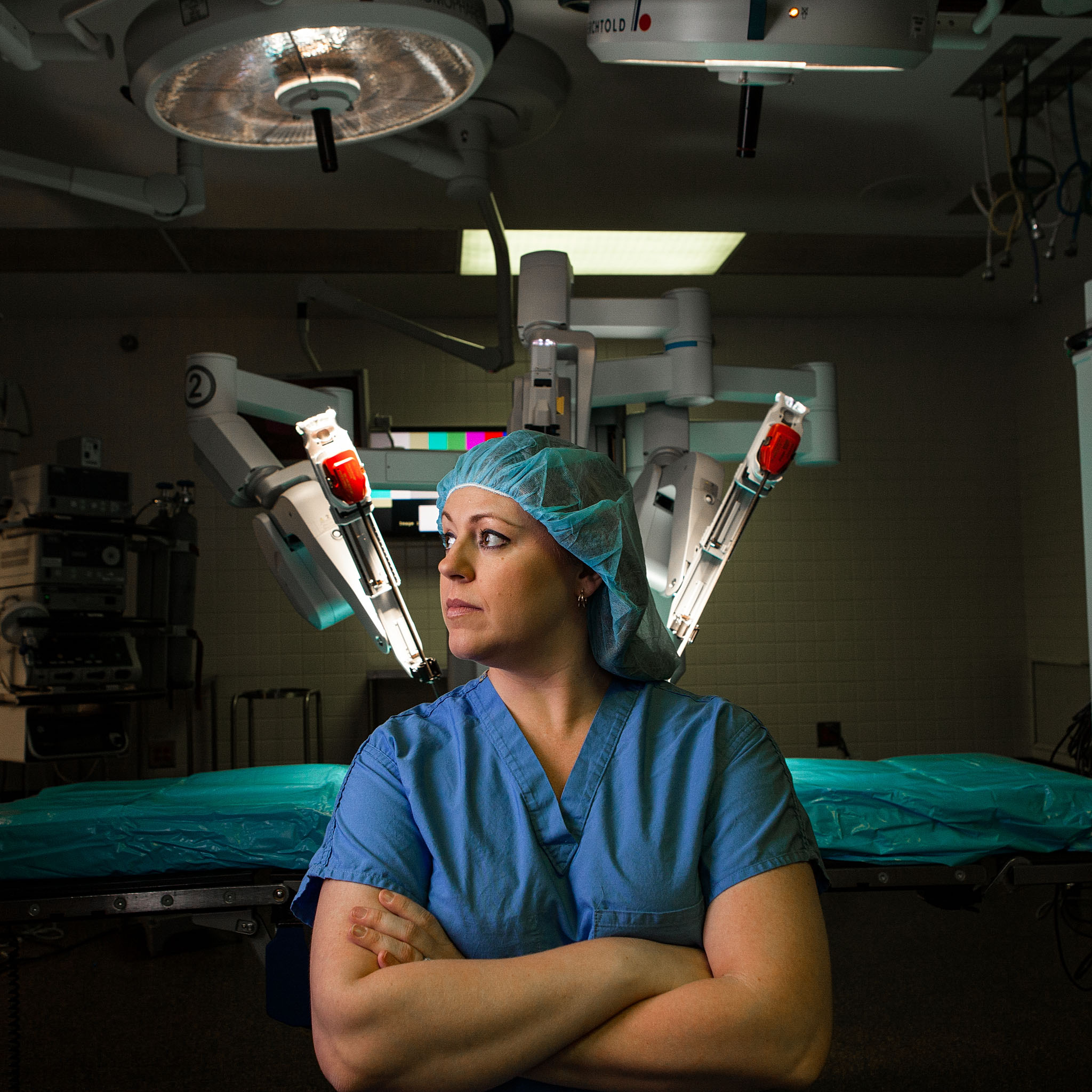 Surgical nurse in blue scrubs and cap with arms crossed in an operating room with a robotic surgical system