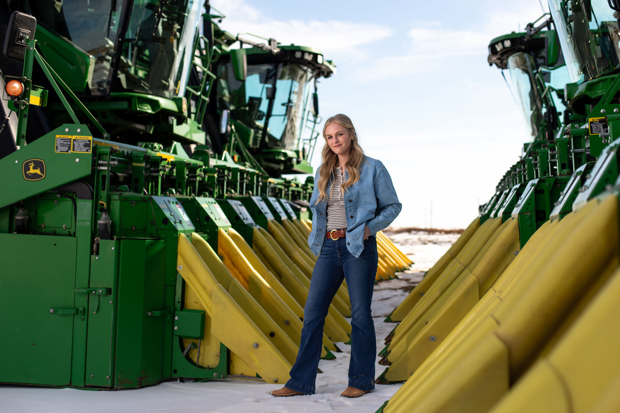 Woman in denim standing between the green and yellow row units of a cotton harvester