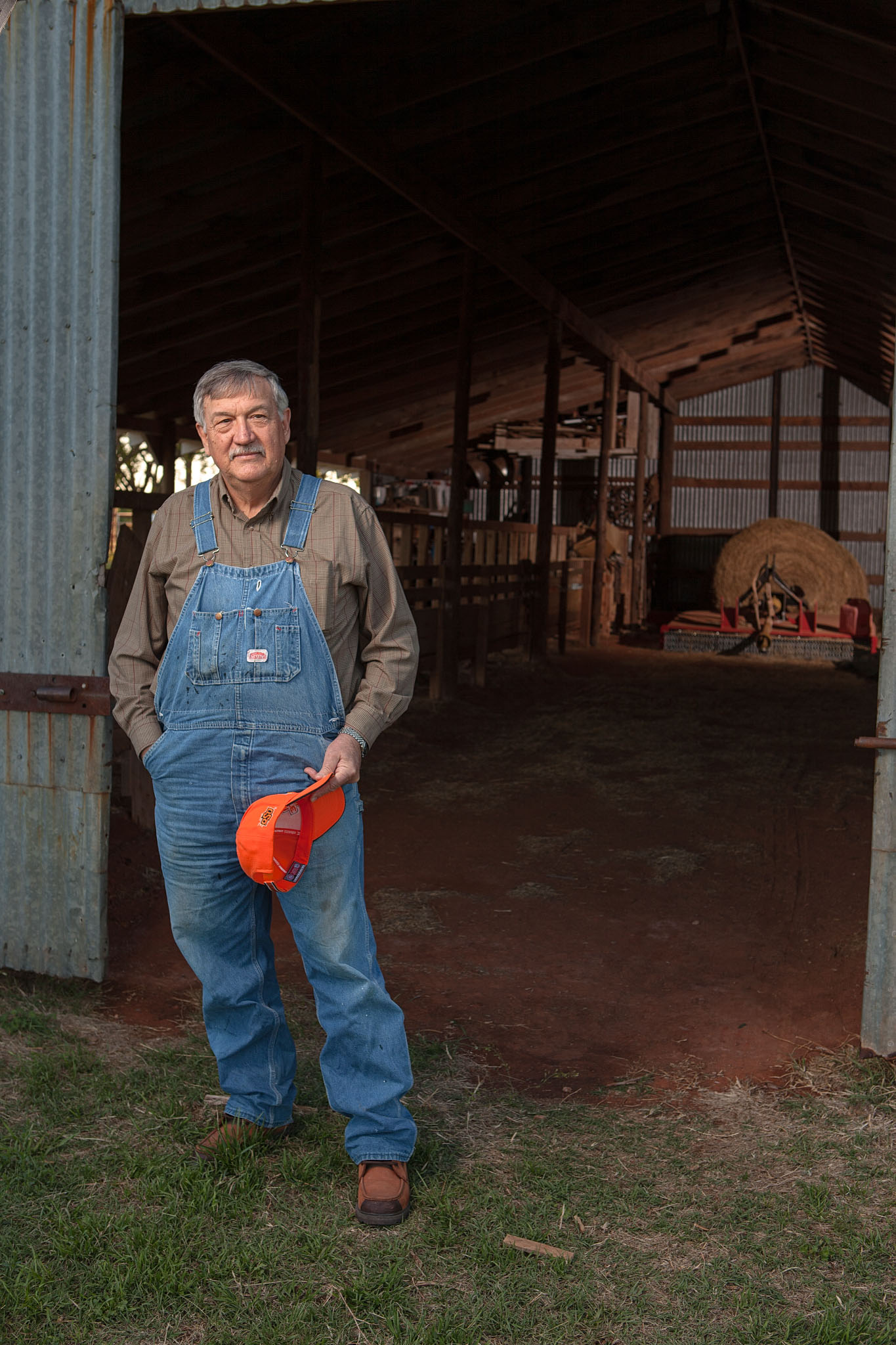 Older man in denim overalls holding an orange cap in a barn doorway