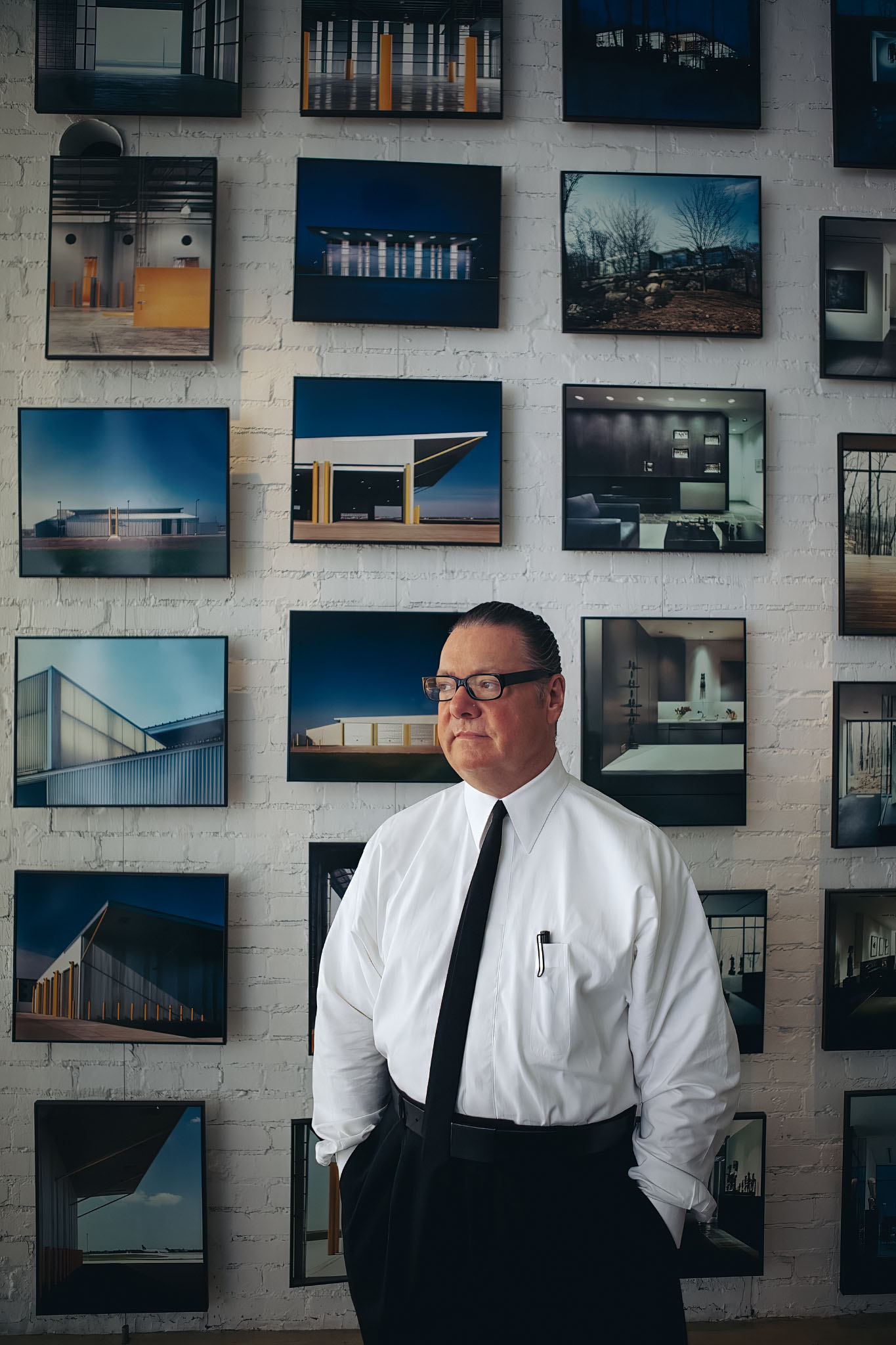 Man in a white shirt and black tie standing before a wall of framed architecture photographs