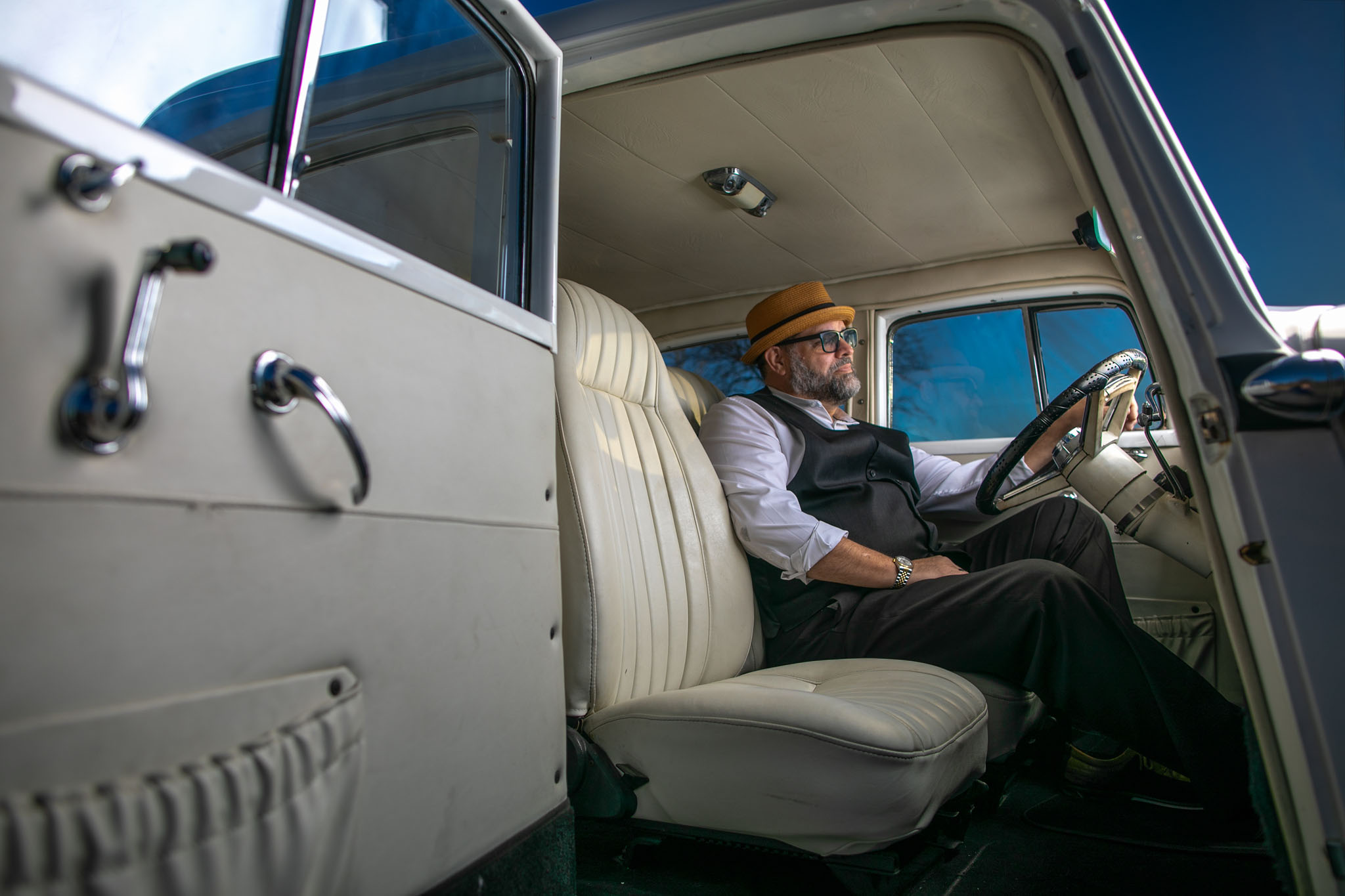 Older man in a hat seated behind the wheel of a vintage car with a cream interior