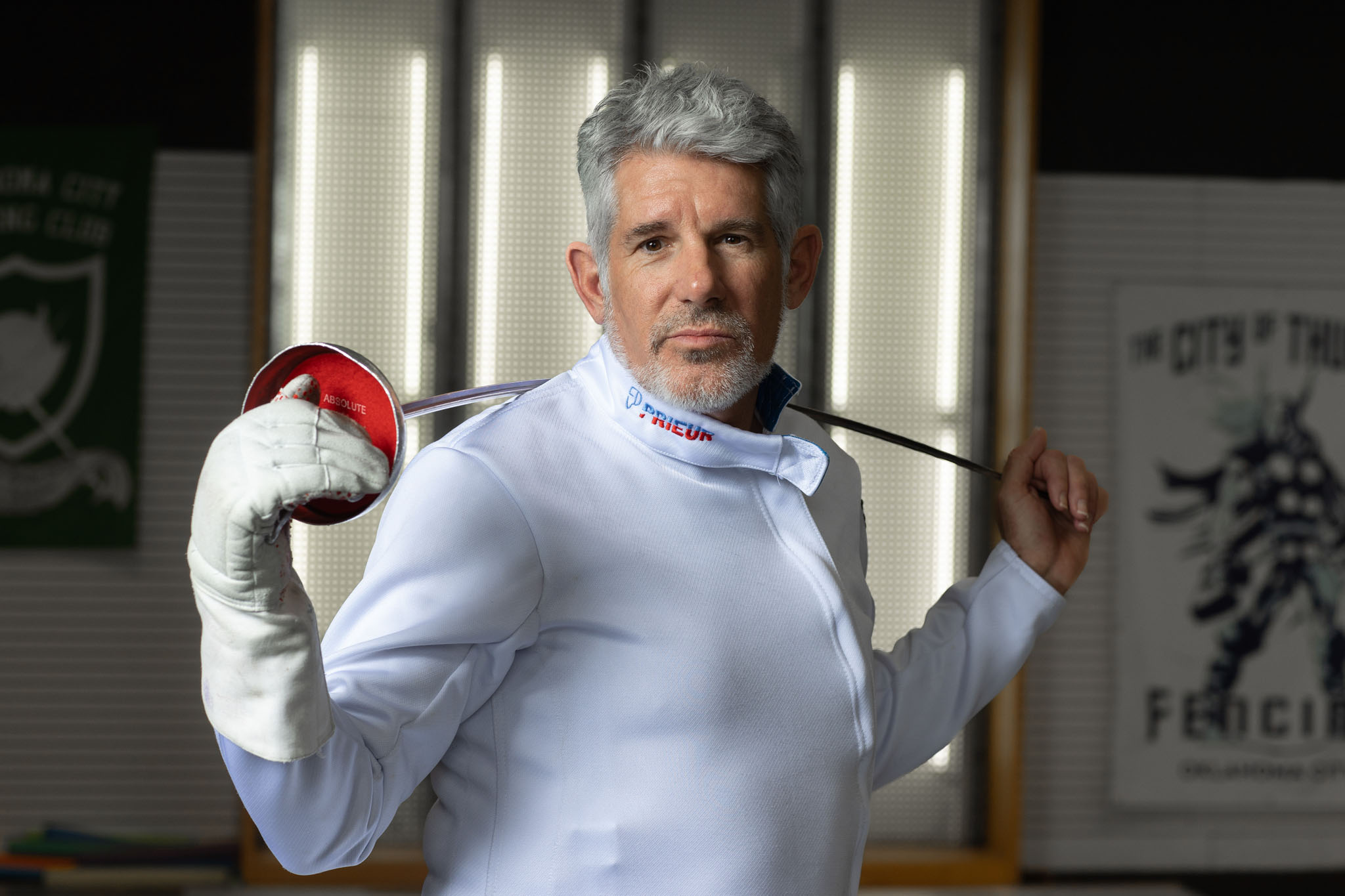 Fencer in a white fencing jacket holding a foil over his shoulder in a fencing club