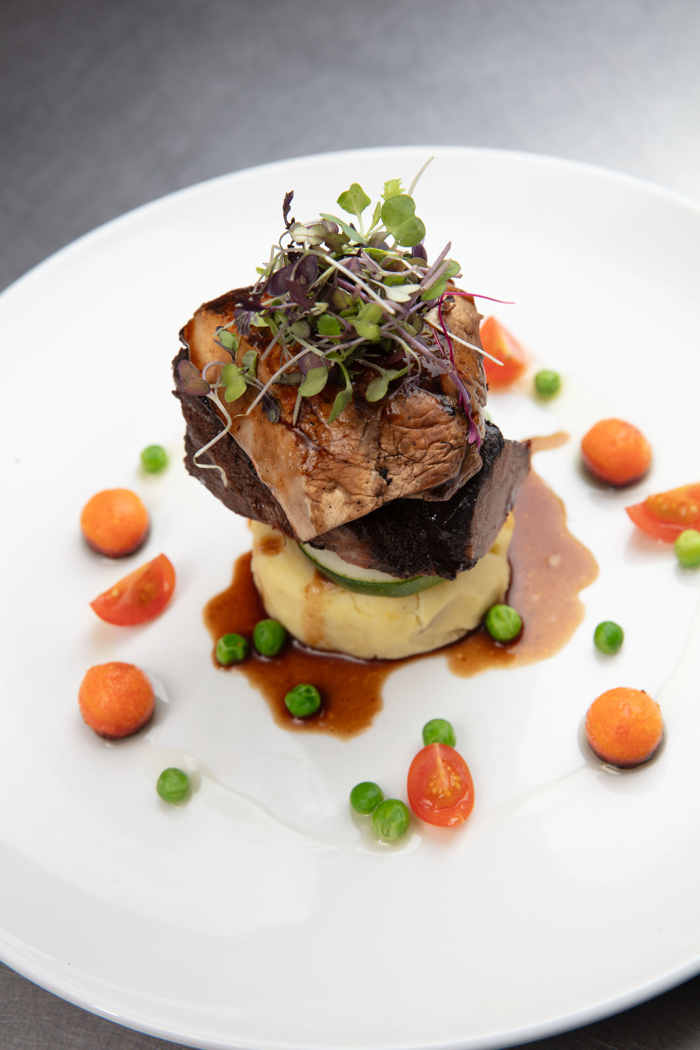 Plated stack of seared meat over mashed potatoes topped with microgreens, surrounded by jus, peas, carrot rounds and cherry tomato halves on a white plate