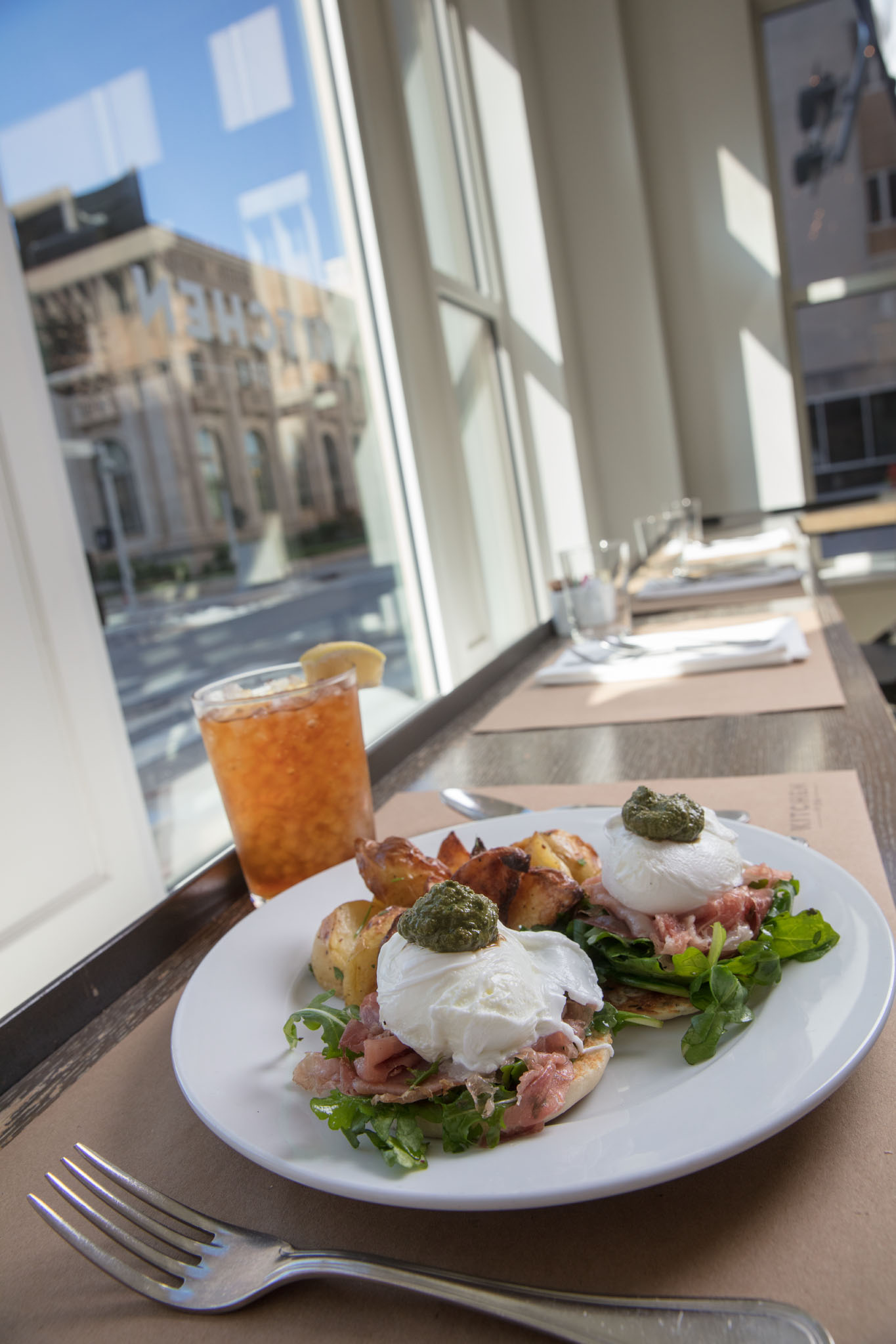 Poached eggs topped with pesto over prosciutto and greens, with roasted potatoes and an iced tea, on a sunlit table beside a tall window