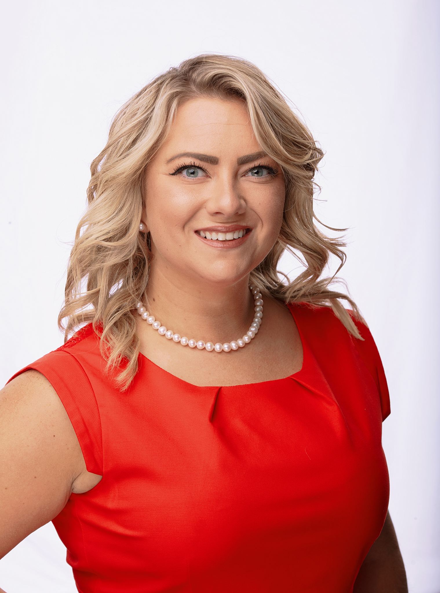 Studio headshot of a smiling woman with wavy blonde hair in a red dress and pearl necklace on a white backdrop