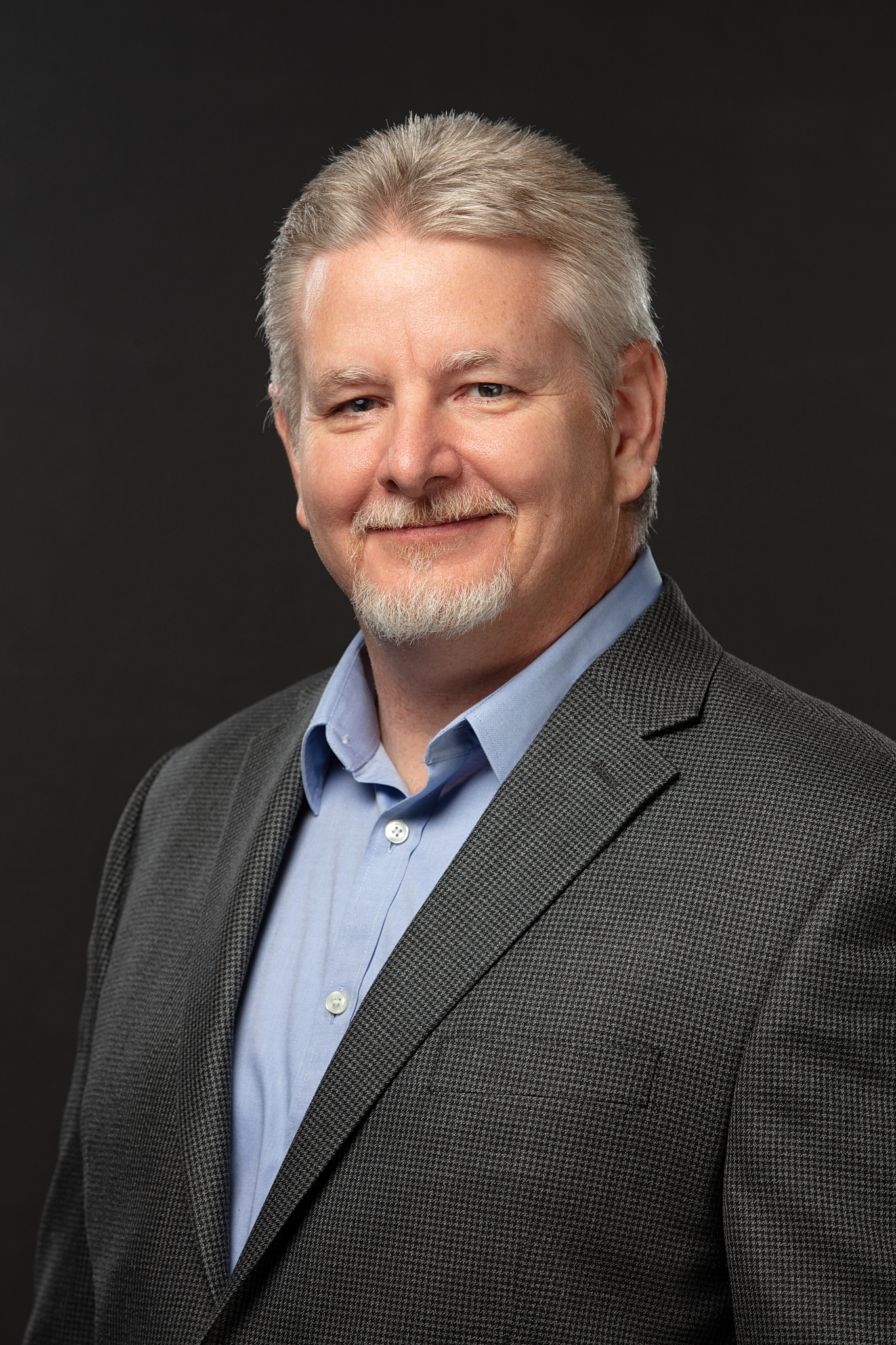 Executive headshot of a man with gray hair in a gray sport coat and light blue shirt on a black backdrop