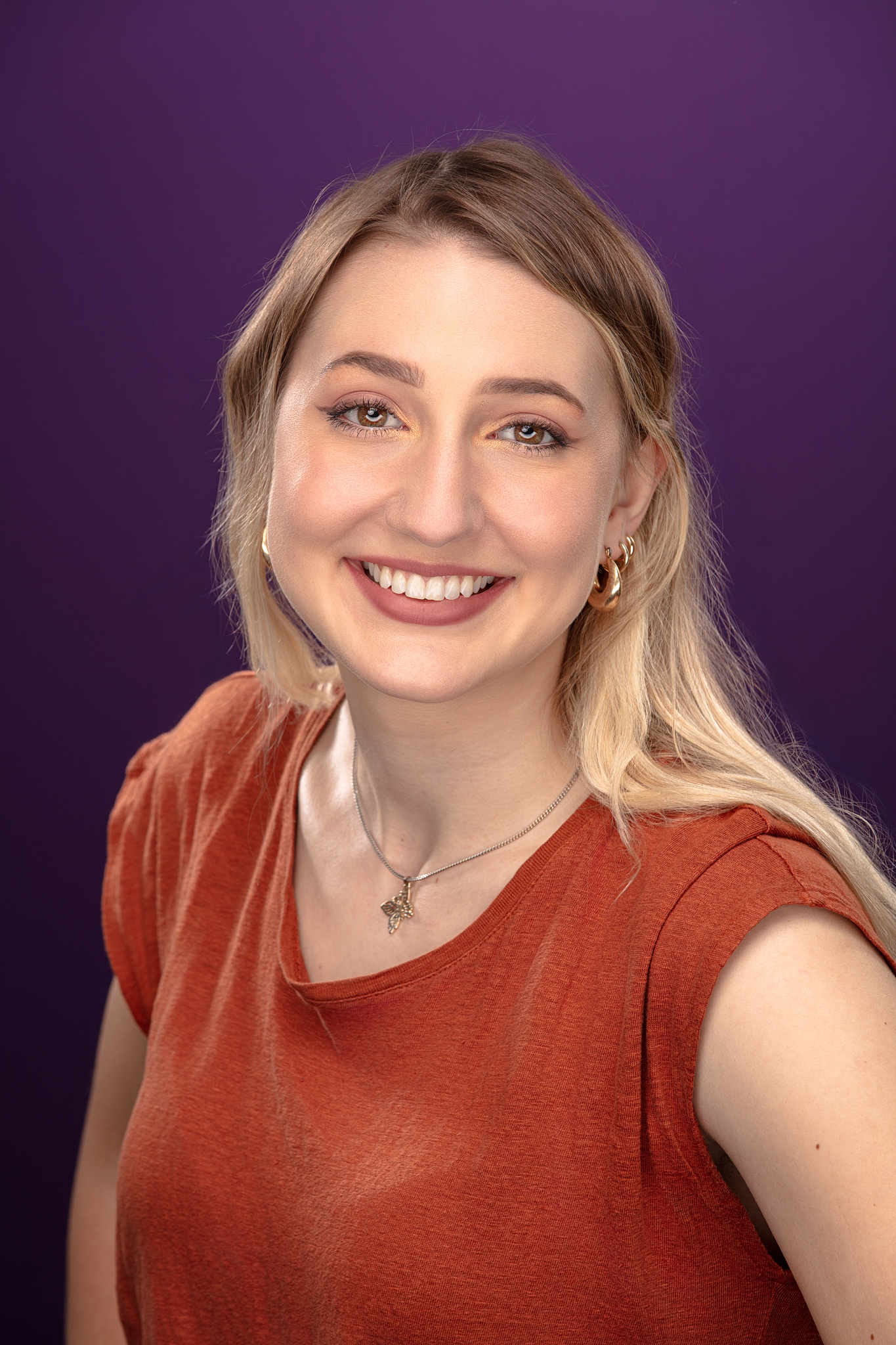 Studio headshot of a smiling young woman in a rust top against a purple backdrop