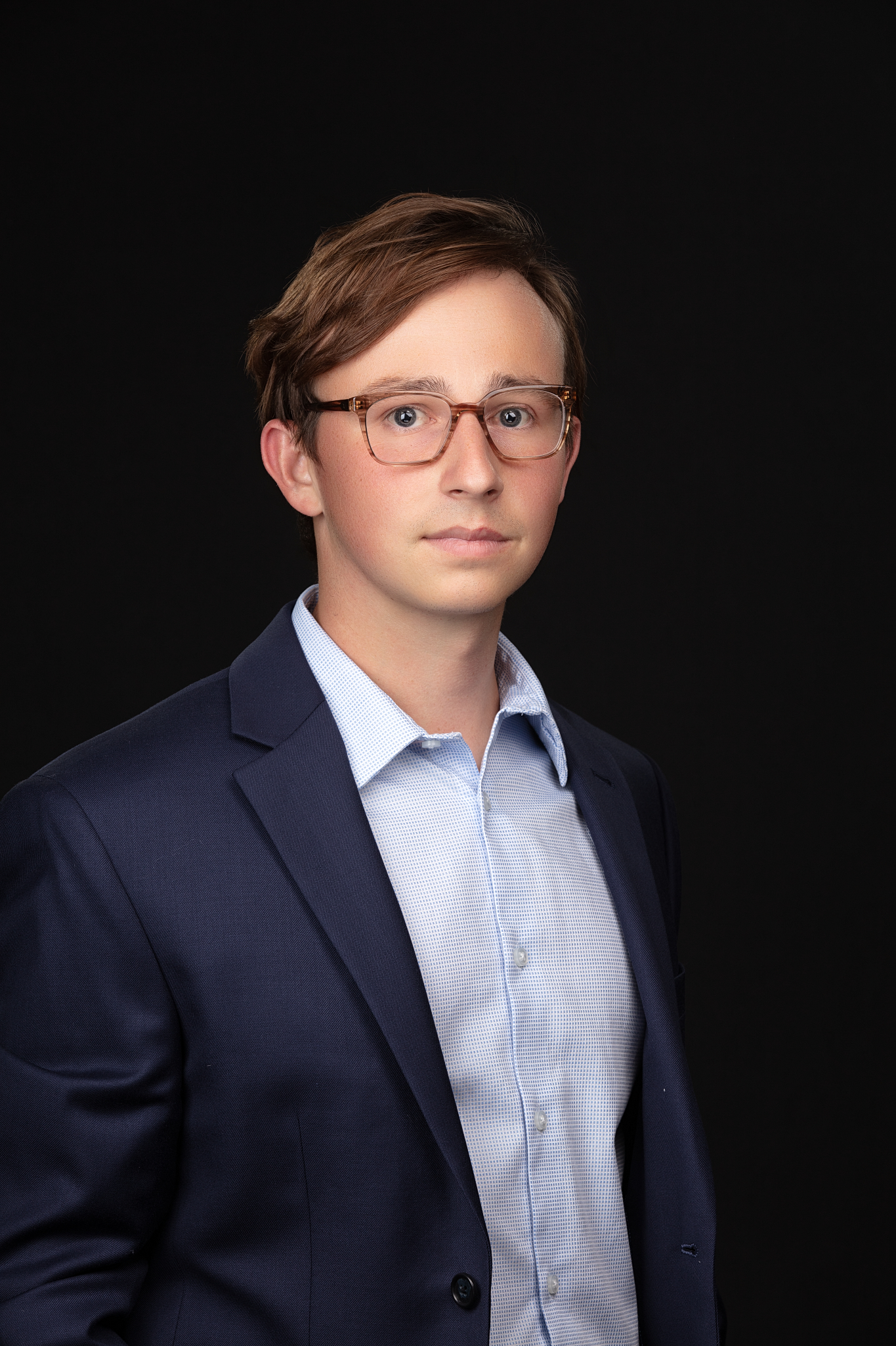 Business headshot of a young man in glasses, navy suit jacket and light blue shirt on a black backdrop