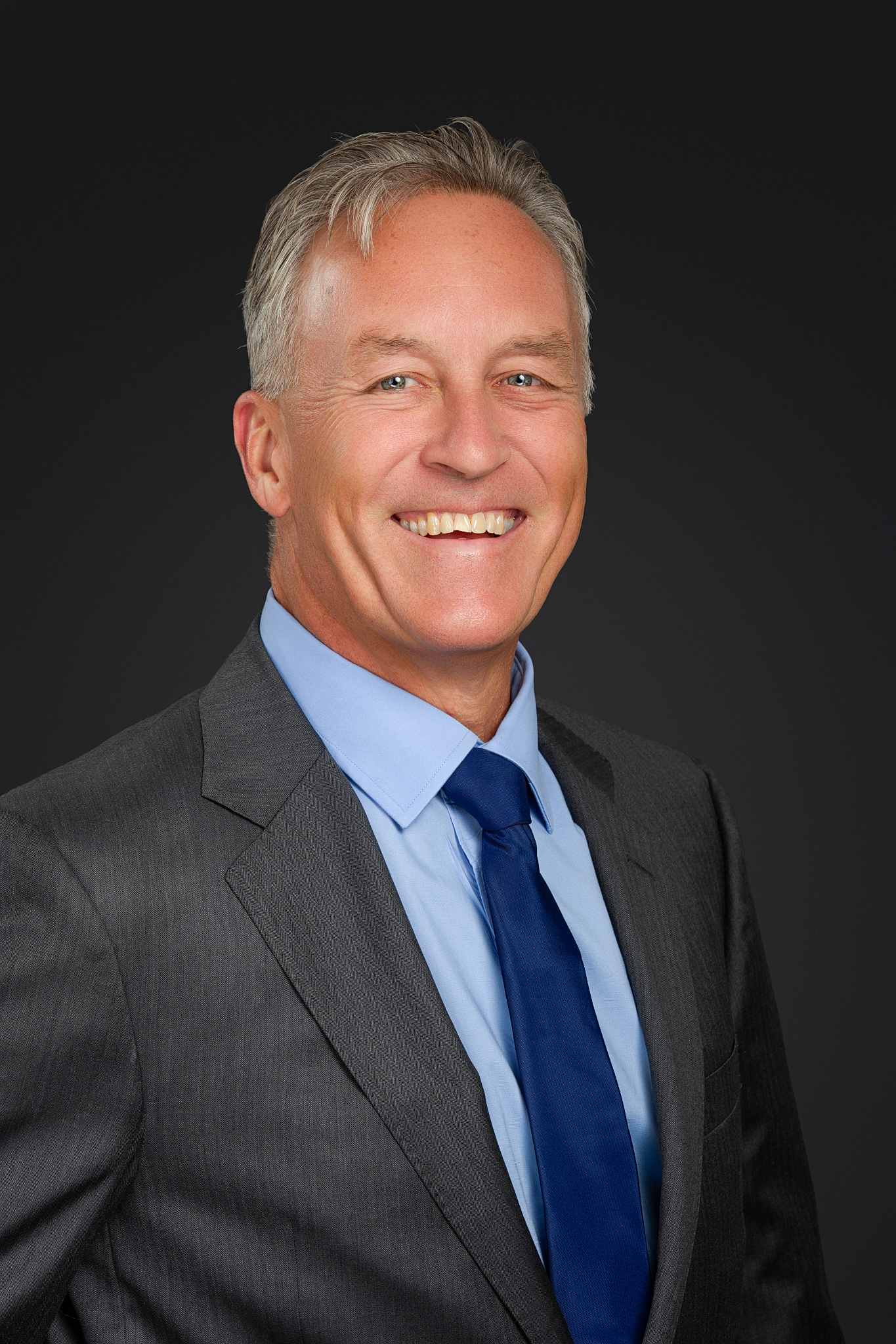 Executive headshot of a smiling man in a gray suit and navy tie on a dark gray backdrop