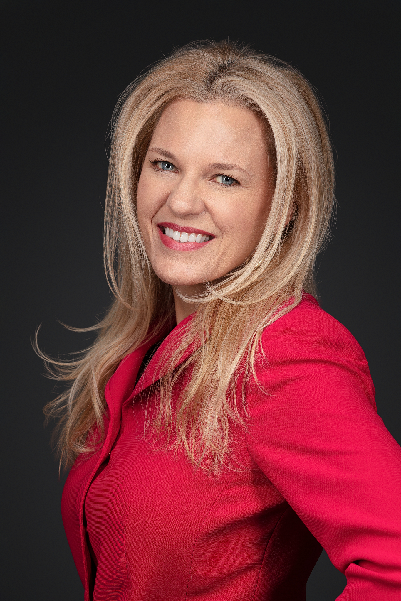 Professional headshot of a smiling woman with long blonde hair in a red blazer on a dark backdrop
