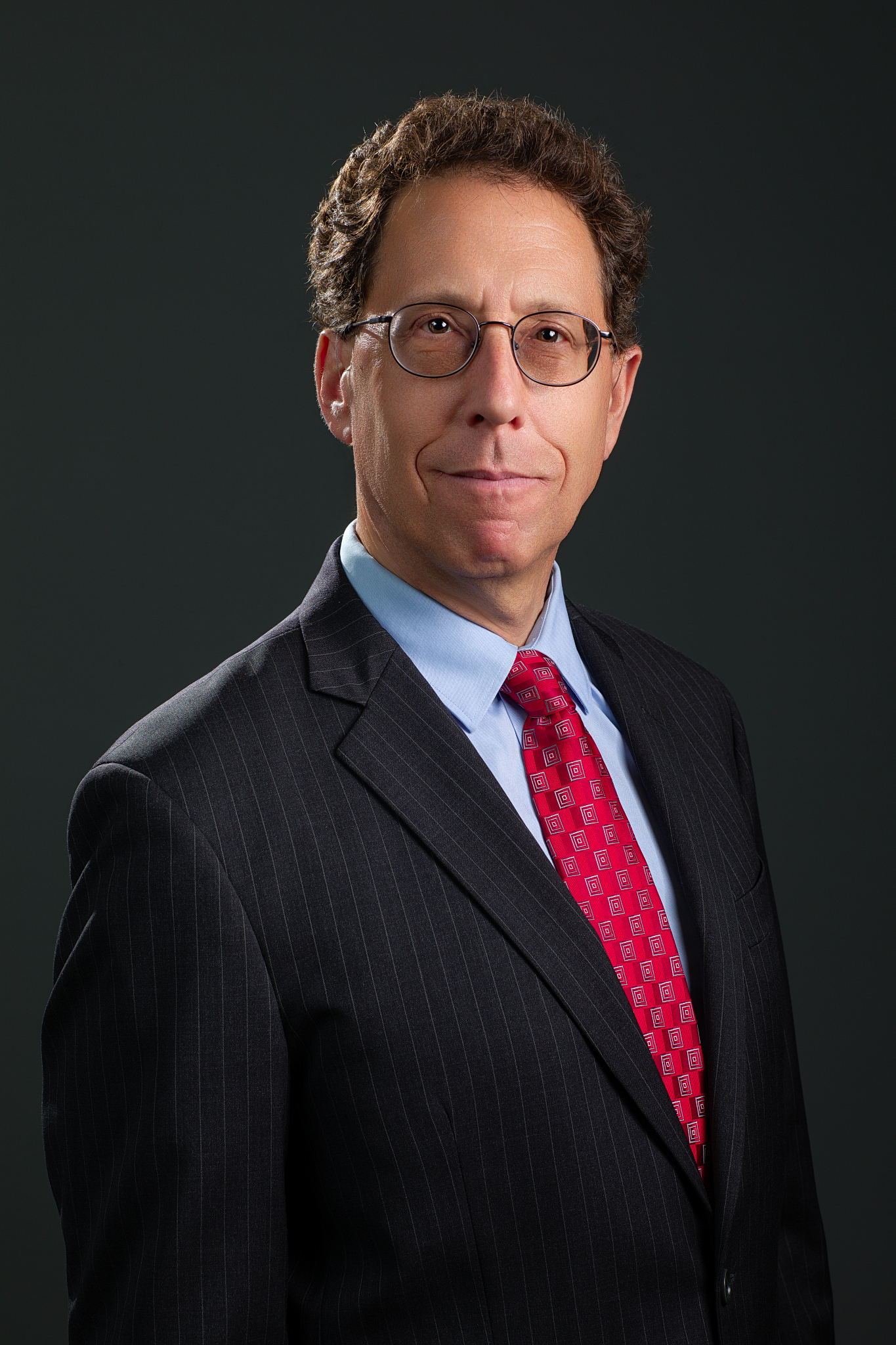 Executive headshot of a man in glasses, pinstripe suit and red tie on a dark gray backdrop