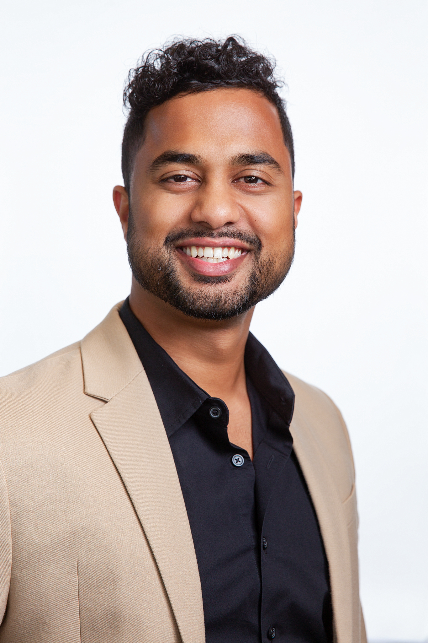 Business headshot of a smiling man in a tan blazer and black shirt on a white backdrop